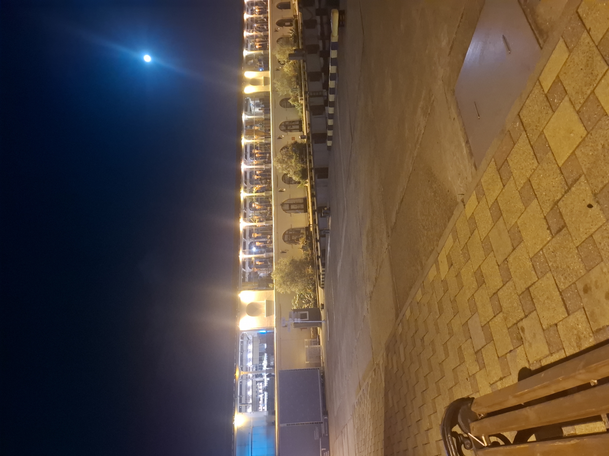 The image depicts an outdoor scene at night, likely in Sochi, Russia. In the background, a long building with a covered walkway is illuminated by numerous lights, revealing tables and chairs, suggesting it may be a restaurant or cafe. The building has arched openings along its facade and is set against a dark night sky where a bright moon is visible. In the foreground, a paved walkway made of yellow and grey rectangular stones leads to a wooden bench. The ground is a mix of paved surfaces and some darker asphalt or concrete. The overall atmosphere is one of a nighttime urban setting, possibly a waterfront or promenade area.