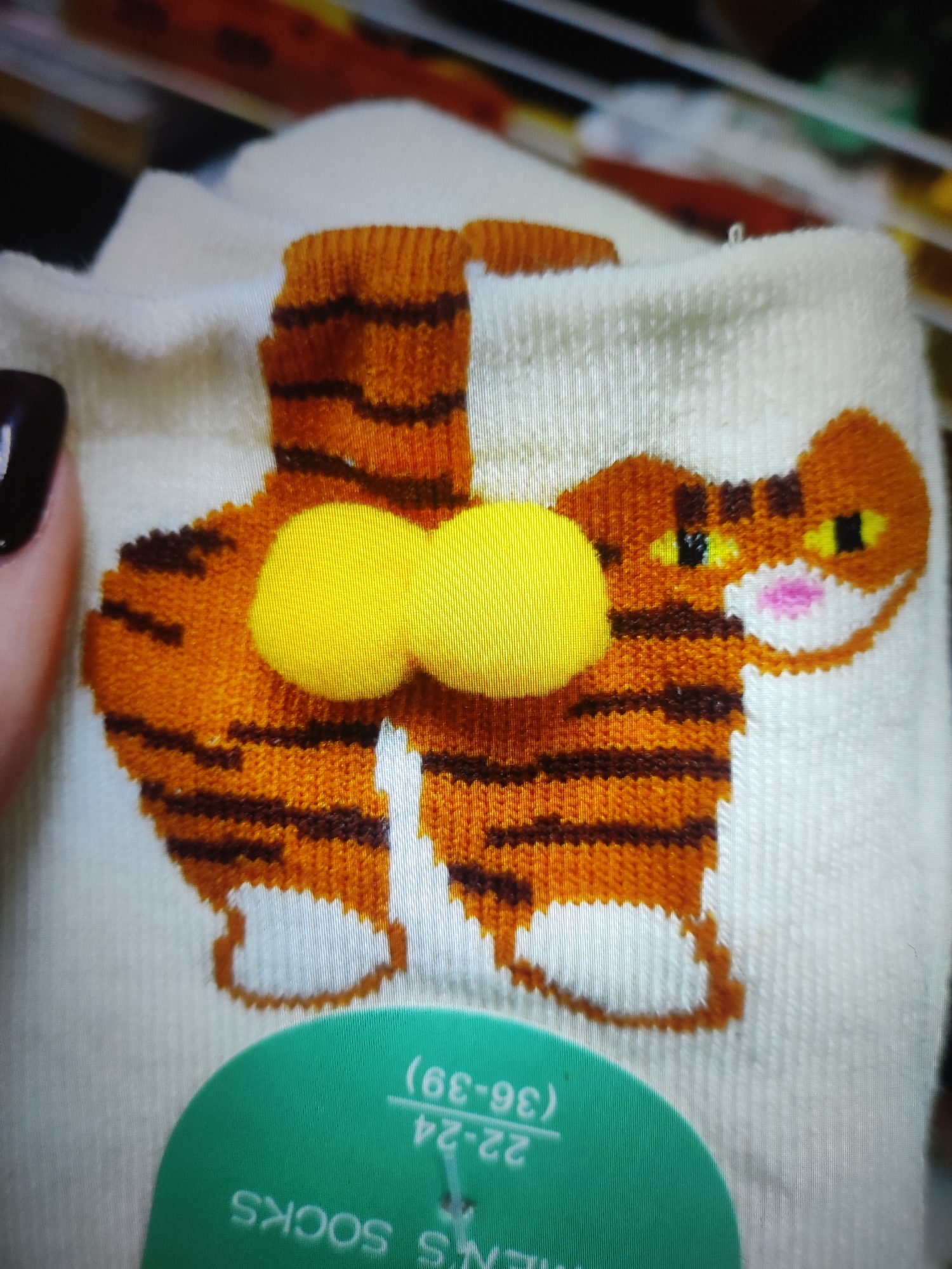 The image shows a close-up of a pair of white socks with a cartoon tiger design. The tiger is depicted from the rear, with its tail pointing upwards and two yellow pom-poms resembling buttocks. The tiger has orange and black stripes. A green tag with white text is visible at the bottom, partially obscuring the lower part of the socks. The text on the tag appears to be in English, with "MEN'S SOCKS" and a size range including "22-24 (36-39)". The background is blurred and appears to be a retail environment, possibly a store shelf with other items. The lighting suggests an indoor setting. There is no visible indication of the location within the city of Cherkasy, Ukraine.