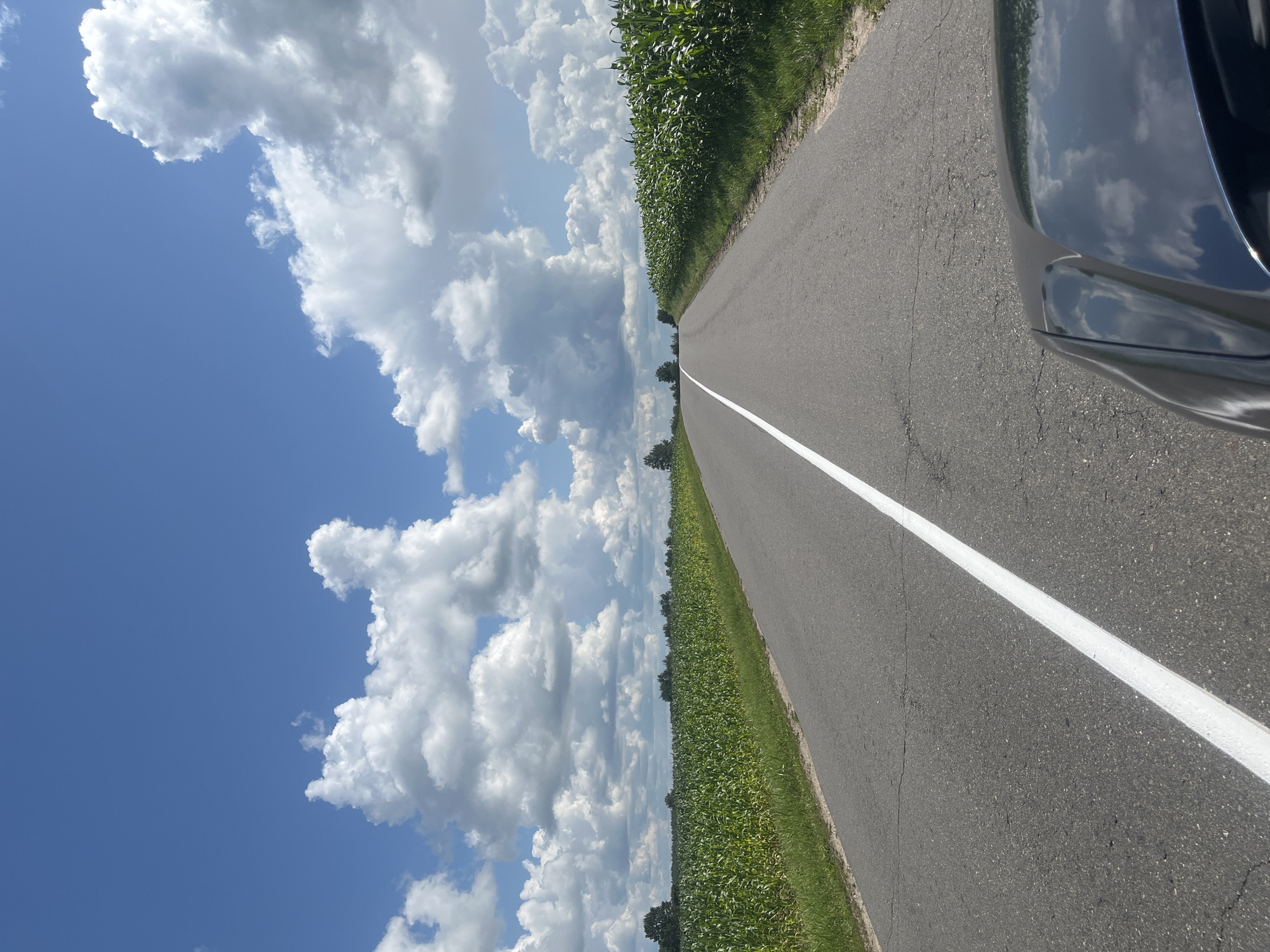 An eye-level, point-of-view shot captures a paved road stretching into the distance under a blue sky with scattered white cumulus clouds. The road, marked with a white center line, is flanked by a lush green field of corn on the right and a similar field on the left, with a line of trees visible in the background. The front of a dark-colored car is visible in the lower right corner of the frame, reflecting the sky and clouds. The scene suggests a rural setting on a clear day.