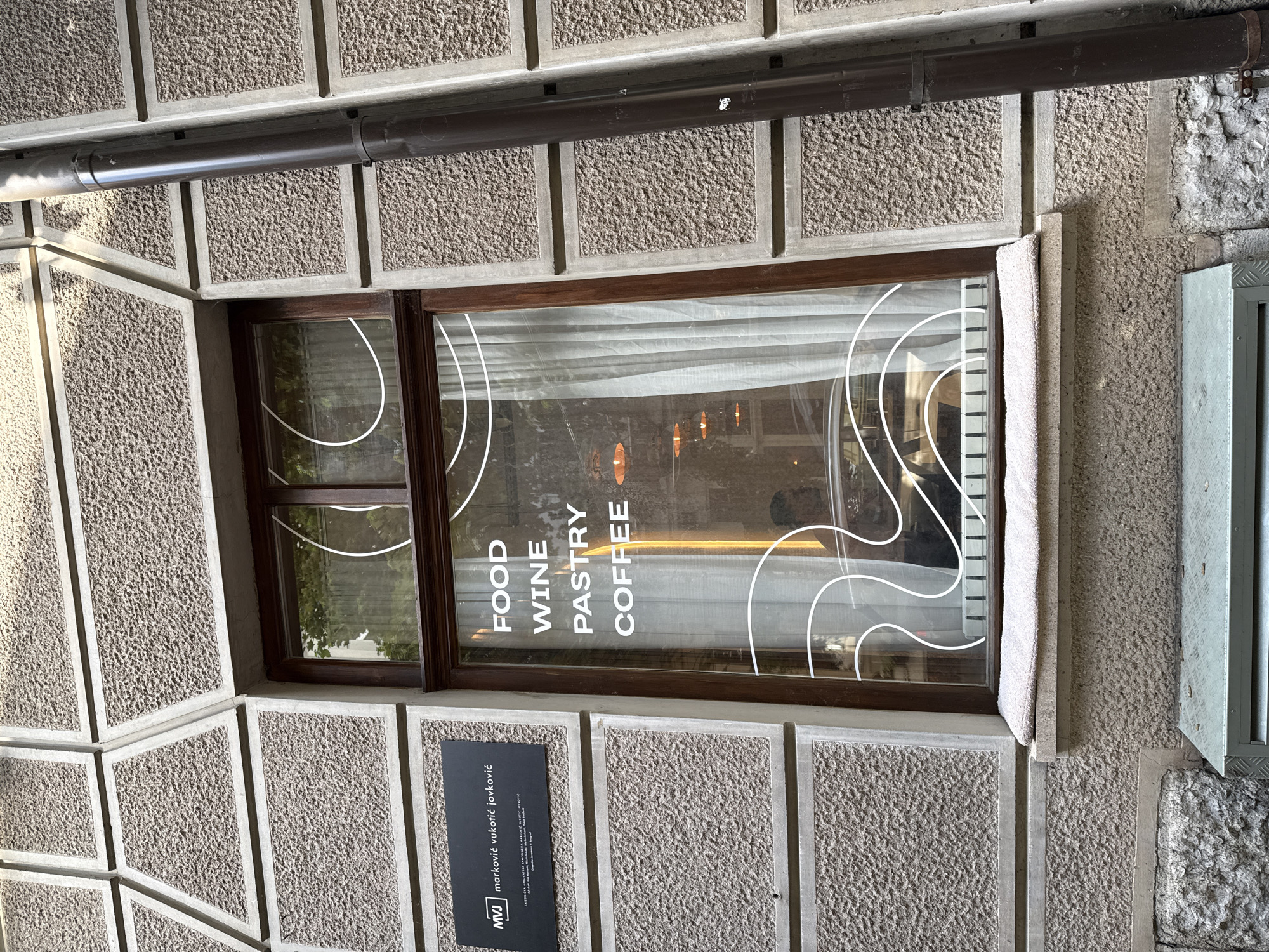 Here is a description of the image:
Exterior shot of a window in a building located in Belgrade, Serbia. The building has a facade made of rectangular, textured concrete blocks. A dark brown rain gutter runs horizontally above the window frame. The window itself has a dark brown frame and consists of two panes. White text is printed on the window: "FOOD WINE PASTRY COFFEE". Additionally, a decorative design featuring stylized white lines is also present on the window. A gray plaque with white text is attached to the wall beneath the window, displaying the name "marković vukotić jovković" along with the acronym "MVJ". The window appears to be part of a cafe or restaurant. The window reveals a glimpse of the interior, including soft lighting and white drapery. To the right of the window, a partially visible metallic structure is affixed to the wall. The lighting is natural, suggesting daylight conditions.