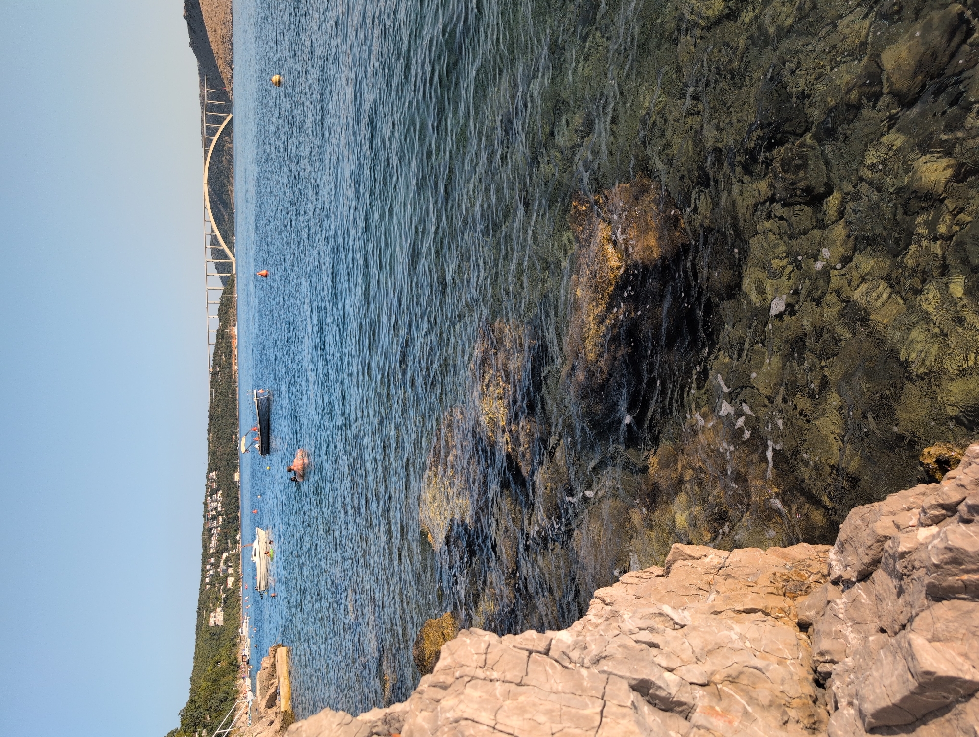 This image depicts a coastal scene in Kraljevica, Croatia, featuring a body of water, a bridge, a shoreline with rocks, and several boats. In the foreground, large, light-brown rocks with visible cracks are prominent. The water in the mid-ground is clear, allowing the rocky seabed to be seen. Several boats are scattered across the water, including a white motorboat and a darker boat. Two individuals are visible in the water; one appears to be swimming, while another is floating on a tube. In the background, a long, arched bridge spans across the water, connecting two landmasses. The shoreline on the left is lined with trees and buildings. The sky is clear and blue, indicating a sunny day.