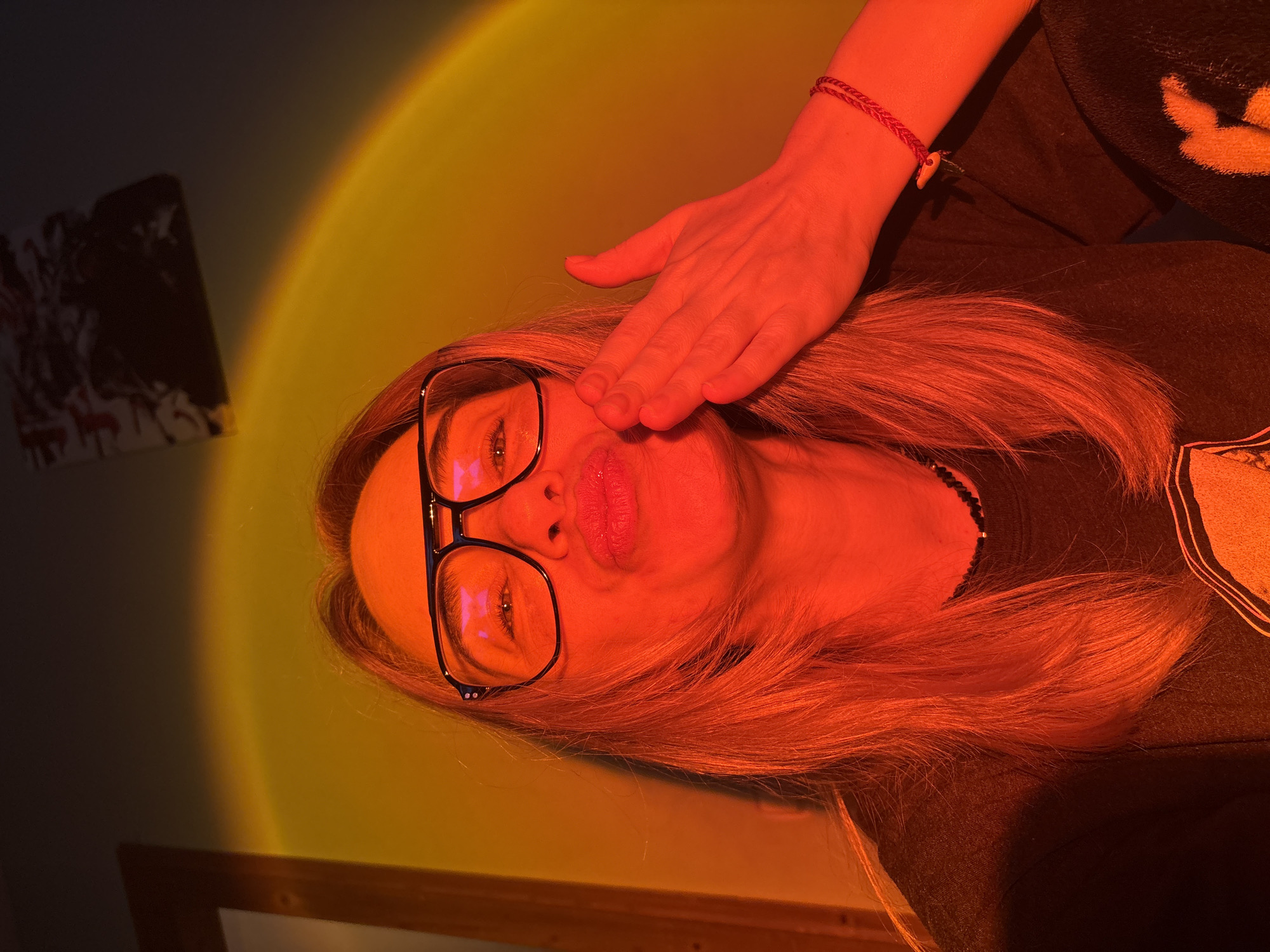 A woman is depicted in an indoor setting, illuminated by a prominent circular light projection that casts warm yellow, orange, and red tones across the scene. Her long, blonde or light brown hair flows around her head. She wears black-framed eyeglasses, through which her eyes are visible, reflecting the colored light. Her right hand is placed over her lower face, covering her nose and part of her mouth, leaving her eyes and forehead exposed. Her lips are slightly parted, suggesting a pout or kissy face. On her right wrist, she wears a red braided bracelet adorned with a small, light-colored charm that resembles a tooth or claw. She appears to be wearing a dark-colored top. In the background, to the left, a framed picture or artwork with dark patterns hangs on the wall, partially in shadow. A wooden surface is visible at the bottom edge of the image. The strong, artificial light creates an artistic and warm ambiance, indicating a posed photograph or selfie. No visible text is present.