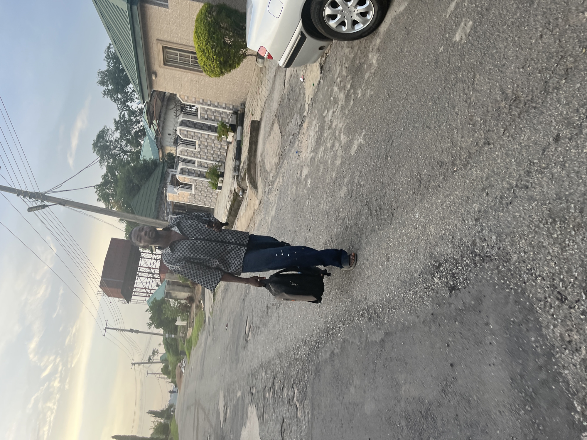 A full-body shot captures a young Black male walking on a paved road in Abuja, Nigeria. He is facing slightly away from the camera, towards the right side of the frame. He wears a patterned short-sleeved shirt, dark blue jeans with metallic embellishments on the side, and black sandals. He carries a dark backpack in his right hand.
The road appears to be in a residential area, with some wear and tear visible on its surface. To the right of the road, a silver car is parked. Behind the car and further down the road, several houses with green roofs and decorative facades are visible. Utility poles with power lines are present throughout the scene, and a large water tank structure stands prominently in the background. The sky is a pale yellow and white, suggesting either early morning or late afternoon light. The overall impression is an everyday scene in a Nigerian urban environment.