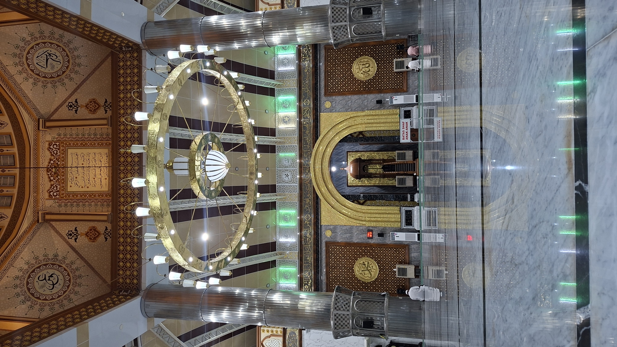 The image depicts the interior of a mosque in Langsa, Indonesia. The central focus is the ornate architecture, including a large, circular chandelier with multiple lights, intricate ceiling decorations featuring Islamic calligraphy and geometric patterns, and gilded archways. Large, fluted metallic columns flank the space. In the foreground, the highly polished marble floor reflects the overhead lighting and architectural elements. Two individuals are visible, seated on the floor in prayer, facing away from the camera. Several air conditioning units are placed along the wall. A sign with red lettering stating "BATAS SHAF ANAK-ANAK" (Children's Prayer Line Limit) is also visible. The overall ambiance is one of grandeur and spiritual serenity.