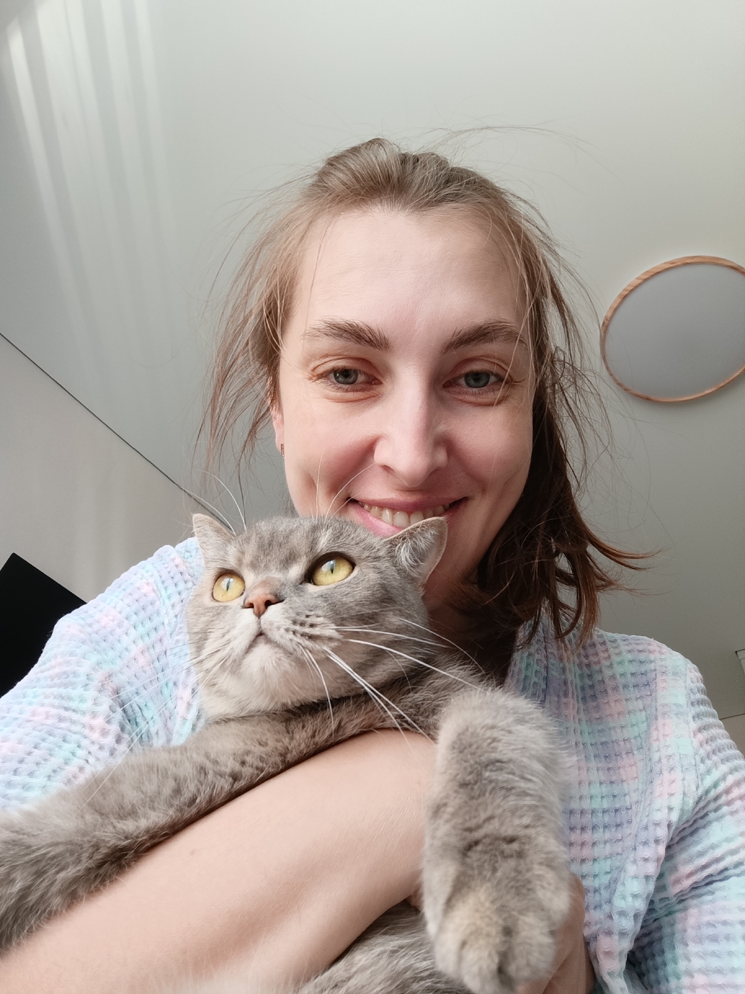 A woman with brown hair and blue eyes smiles at the camera while holding a grey tabby cat. The woman is wearing a light-colored, textured robe. The cat has yellow eyes and looks upwards. They appear to be indoors, with a white ceiling and a round light fixture visible in the upper right corner. The location is likely in Chaikovsky, Russia.