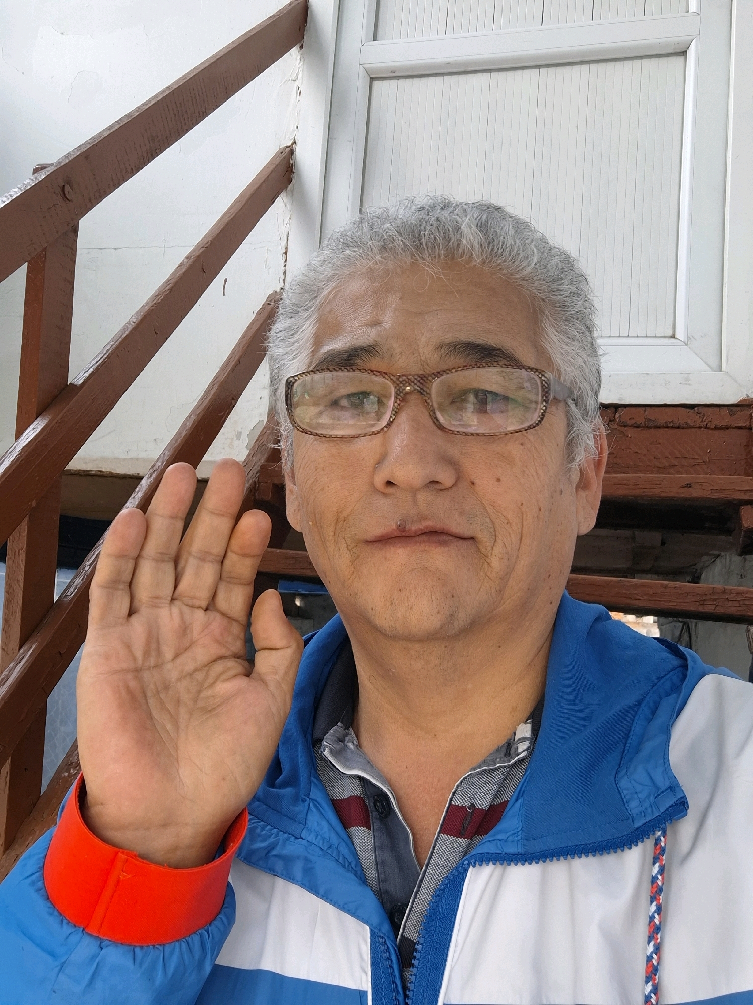 An adult male with graying hair, wearing glasses, and a blue and white jacket with a red cuff is shown from the chest up. He is holding his left hand up, palm facing forward. The man appears to be standing on outdoor stairs. The background shows wooden railings and a white wall. The image is a selfie.