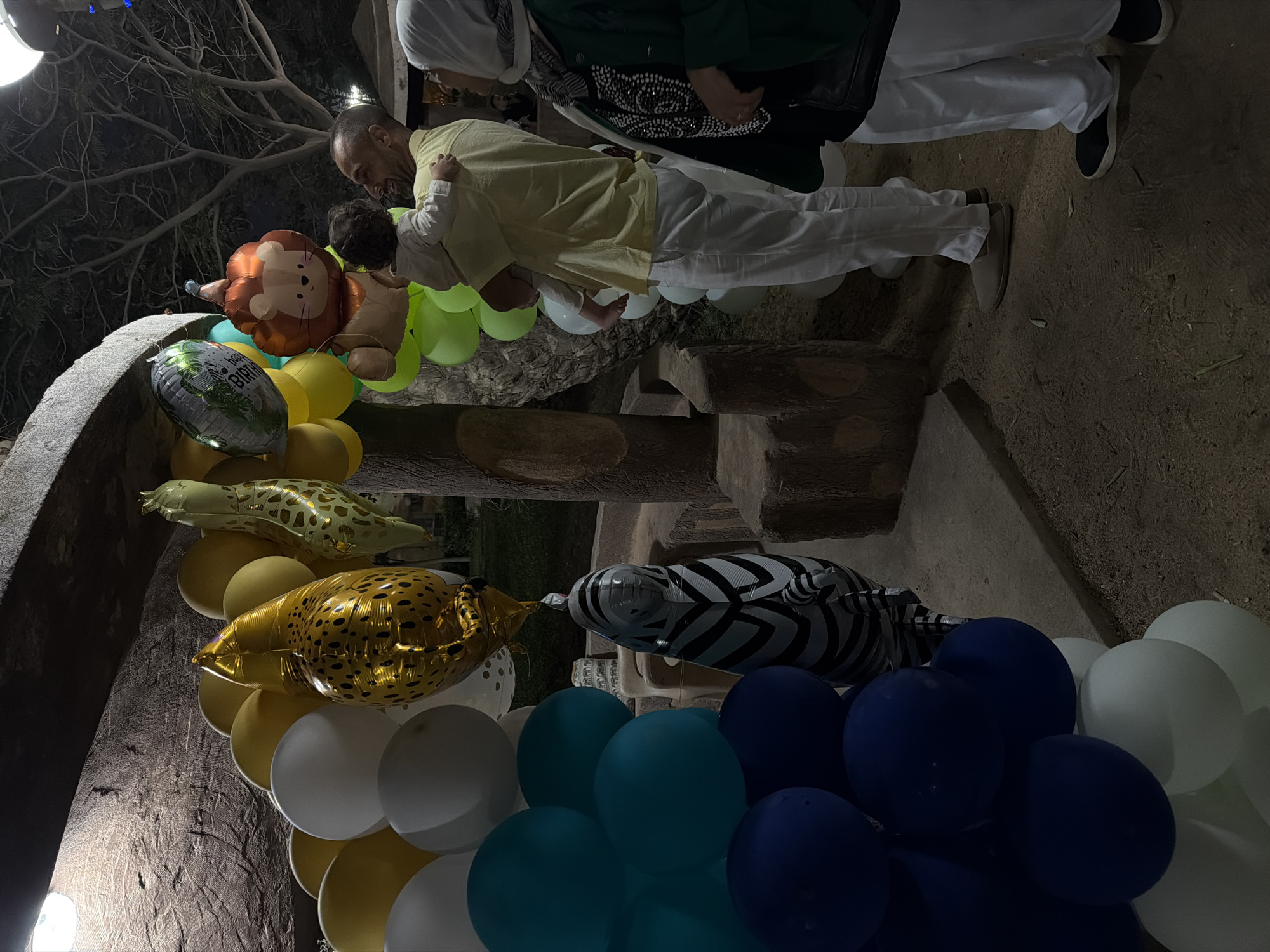 In the city of 6th of October, Egypt, a group of people stand near a collection of animal-themed balloons. The balloons, arranged in a festive display, include a gold cheetah-print balloon, a giraffe-patterned balloon, a lion-shaped balloon, and a zebra-shaped balloon. A man in a yellow shirt holds a young child in his arms. Another person, wearing a green jacket and a white hijab, stands nearby. The scene appears to be an outdoor celebration, possibly a birthday party, held at night. The ground is sandy, and trees are visible in the background.