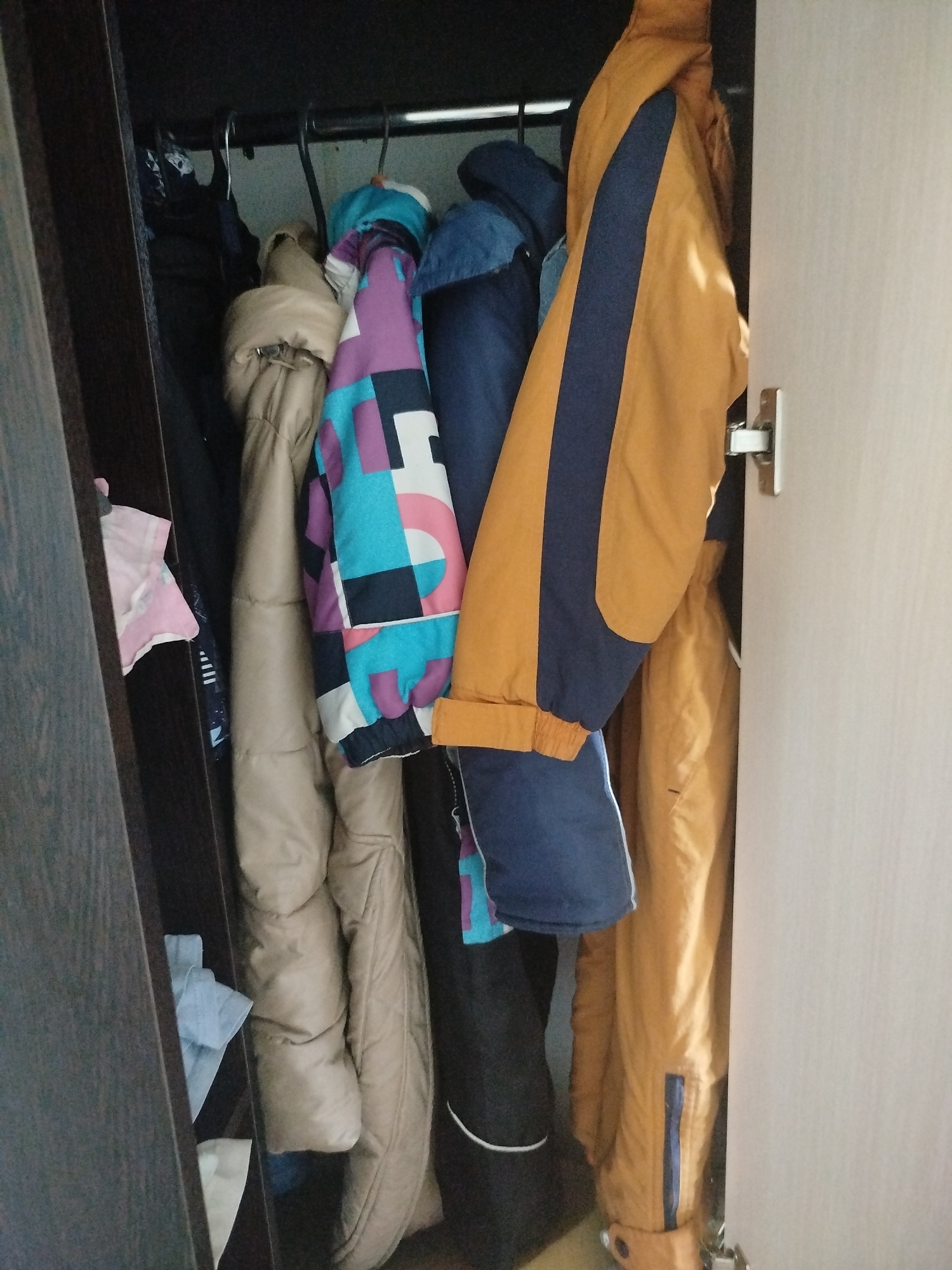 The image shows the inside of a closet, filled with various winter coats and jackets hanging on a rod. The coats are of different colors and patterns, including a beige puffer jacket, a multi-colored jacket with geometric shapes in teal, purple, pink, and black, and an orange jacket with navy blue stripes. Some of the jackets appear to be made of waterproof material, suggesting they are suitable for cold weather. The closet also contains some folded clothing items in a lower section. The overall impression is of a practical storage space for outerwear. The location of Zavodoukovsk, Russia, does not have any visual cues in the image itself, but it provides a geographical context for the type of clothing shown. The image is likely taken indoors, in a residential setting.