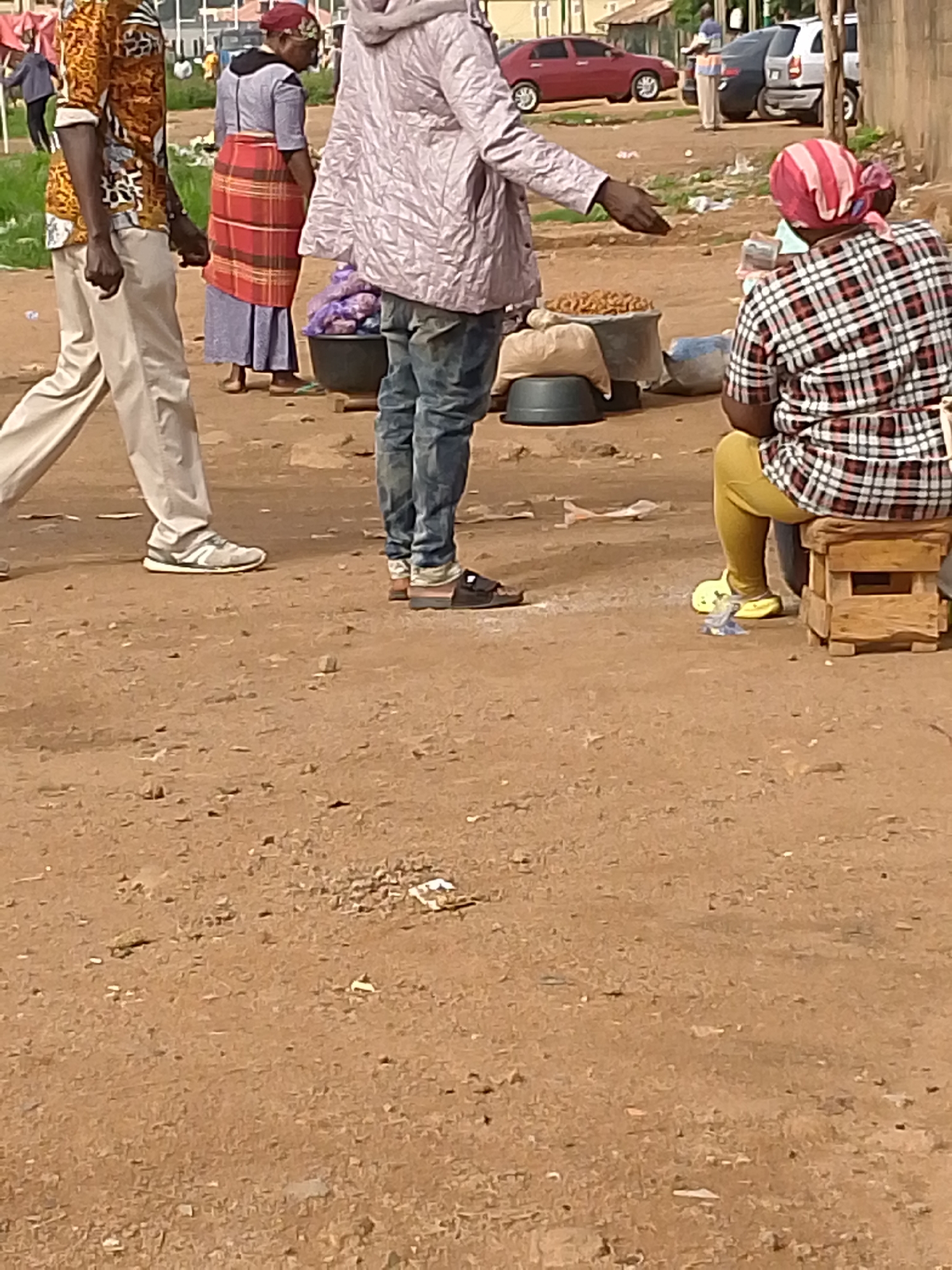A street market scene is depicted in Bukuru, Nigeria. In the foreground, three individuals are visible. On the left, a person wearing a patterned shirt and light-colored pants walks away from the camera. In the center, another person in a light-colored jacket and blue jeans is extending their left hand, palm up, towards the right. To the right, a person wearing a checkered shirt and yellow pants sits on a wooden stool, facing away from the camera. In front of the seated person, there are bags of what appear to be goods, and to the left of the seated person, a large black container holds purple items. The ground is composed of brown dirt with scattered small stones and debris. In the background, cars are parked and buildings are visible.