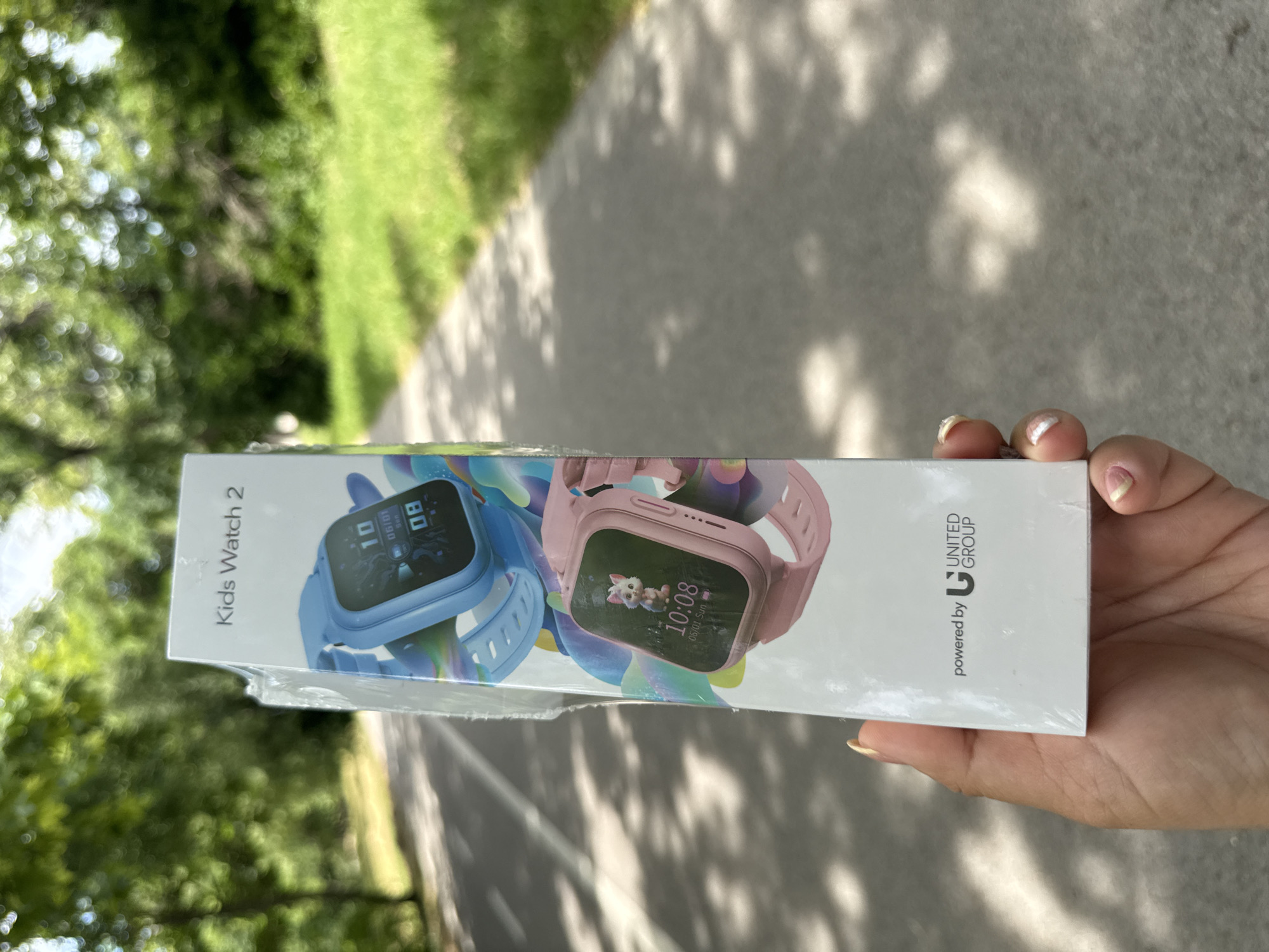 The image shows a person's hand holding a box of "Kids Watch 2". The box features images of two smartwatches, one blue and one pink. The blue watch displays the time "10:08" and the date "06/01 Sun". The pink watch also shows "10:08" and "06/01 Sun". Text on the box reads "Kids Watch 2" and "powered by UNITED GROUP". The background is blurred, but appears to be an outdoor setting with greenery and a paved path, suggesting a park or similar environment. Sunlight is casting shadows on the path. The image is taken from a high angle, looking down at the box and the hand. The person's fingernails are visible and painted.