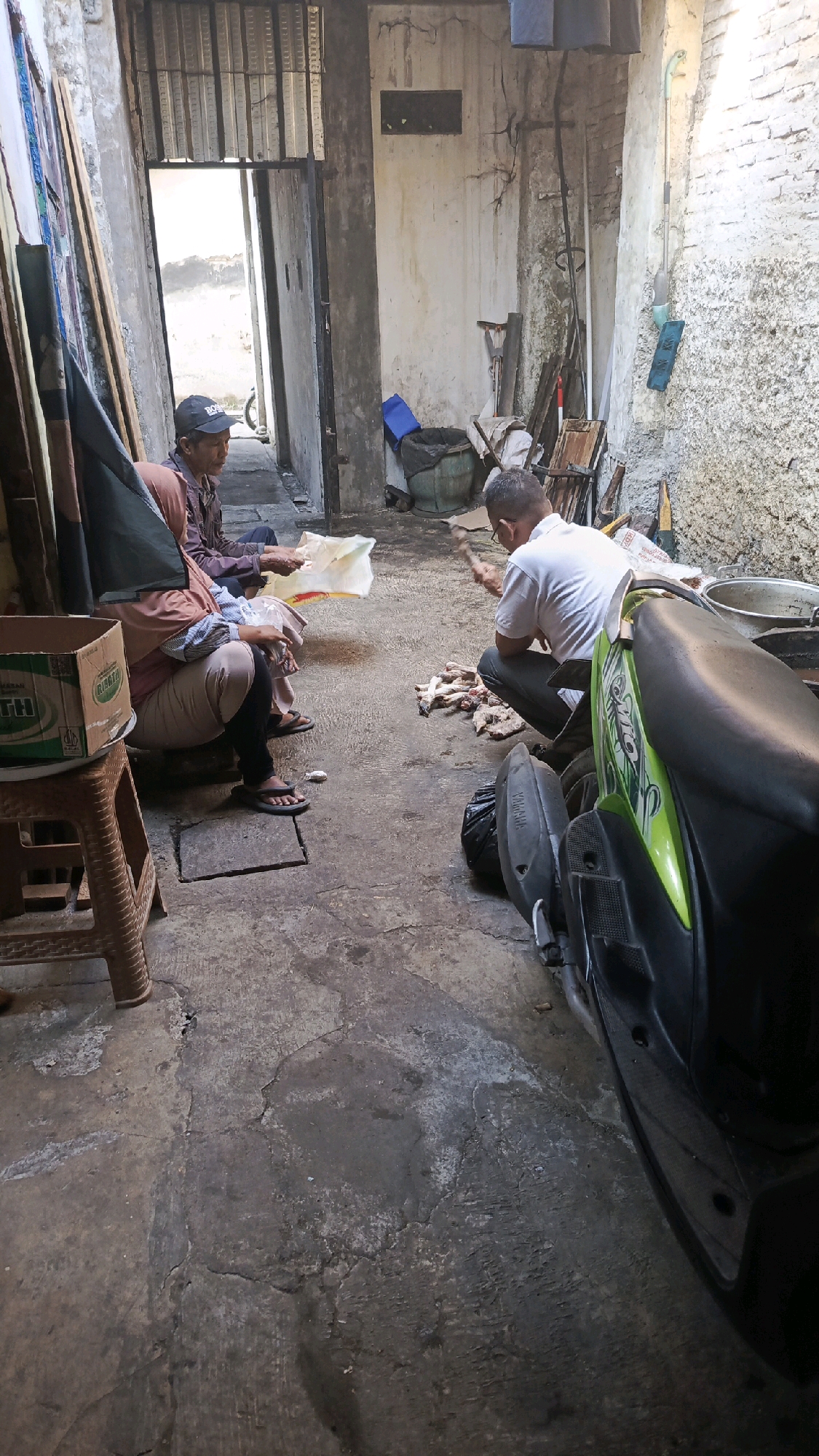 In the city of Pekauman Kulon, Indonesia, an outdoor, open-air, concrete-floored space appears to be a workshop or a service area. Sunlight streams in from an open doorway at the rear, illuminating a gritty, worn environment. A man in a white shirt and dark pants squats on the ground, holding a cleaver and appears to be in the process of butchering a pile of meat. To his left, a woman in a pink headscarf and beige pants is also seated, holding what looks like a plastic bag. Another person, partially obscured, sits to her left. A green motorcycle is parked in the foreground, its front wheel angled towards the left. Various items are scattered around the scene, including a brown cardboard box on a brown plastic stool, a blue plastic object, a green bucket, and other indistinct objects. The walls are rough, textured concrete, with evidence of cracks and wear. The overall impression is one of a functional, possibly informal, area where work is being carried out.