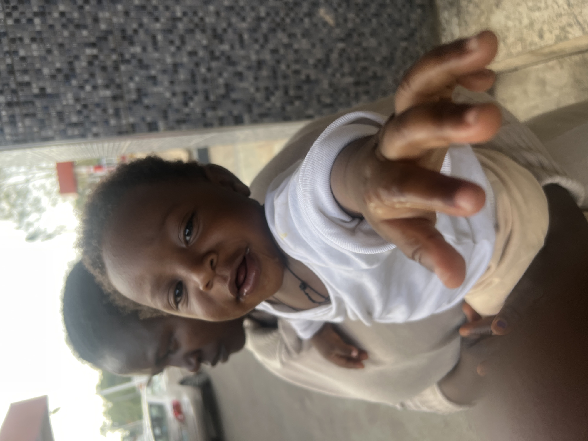 A baby is held up towards the camera by an adult. The baby is wearing a white, long-sleeved shirt and has a necklace. The adult's face is partially visible in the background. In the background, there's a building with black and grey tiled walls. A car is also visible in the background. The location is likely Gura Top, Nigeria.