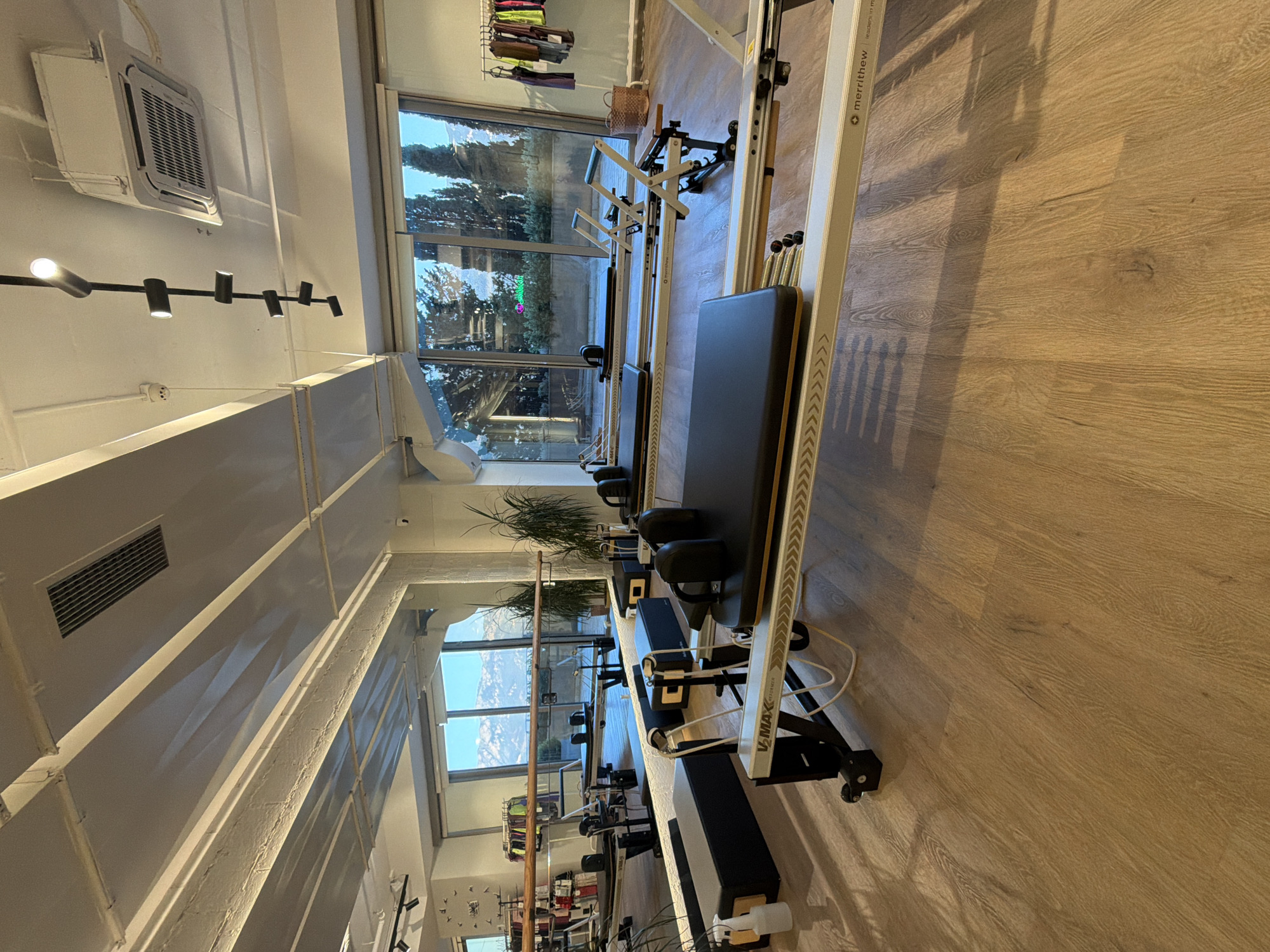 The image shows the interior of a pilates studio. There are multiple pilates reformers, all with black upholstered carriage pads and silver metal frames. The studio has large windows that look out onto a mountainous landscape and trees. The floor is made of light-colored wood. In the background, there is a barre, mirrors, and a rack of athletic clothing. There are no people visible in the image. The studio appears to be in Boreti, Montenegro, given the distinctive mountainous scenery visible through the windows which is characteristic of the region.