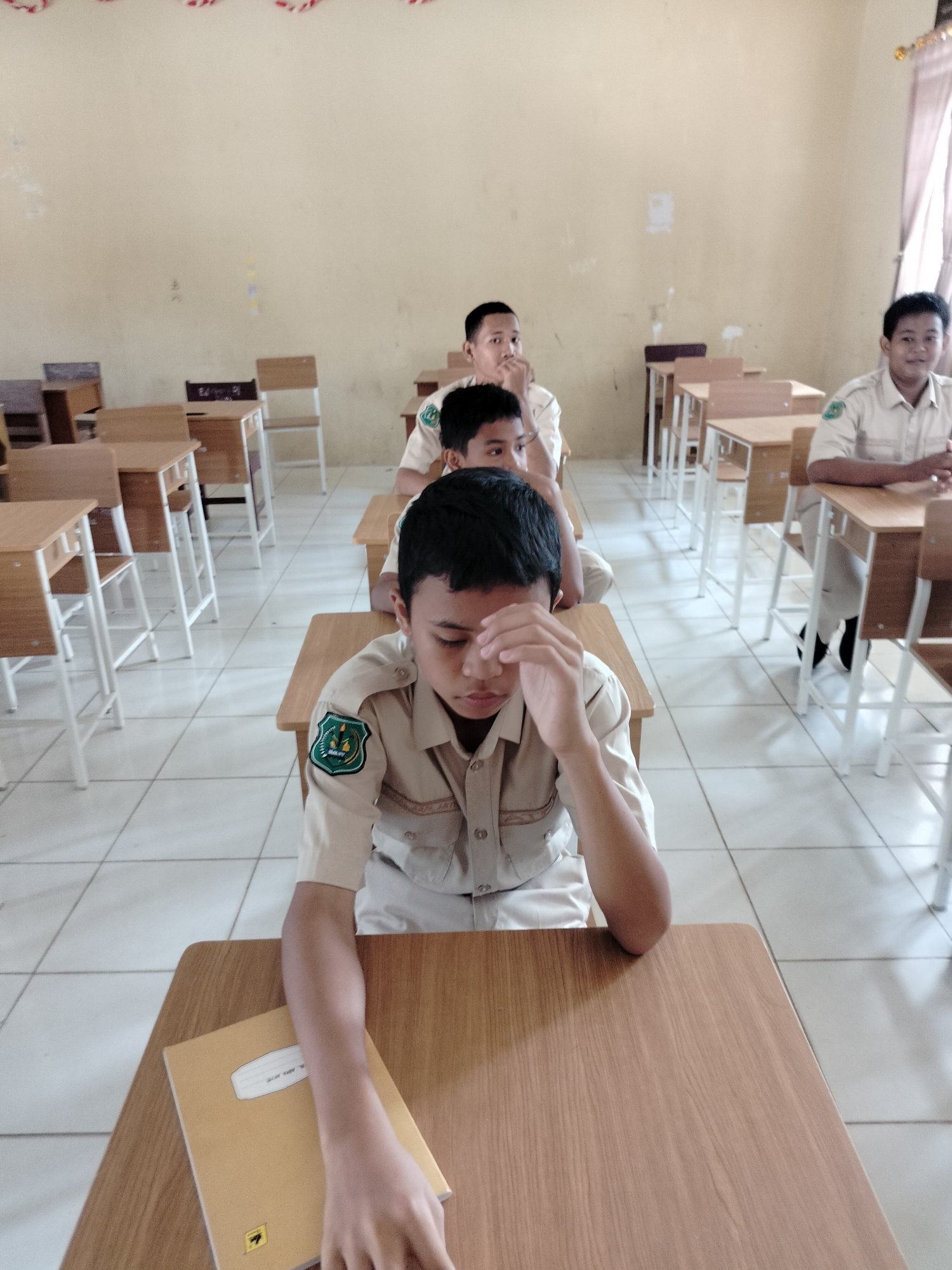 In a classroom in Lawebekung, Indonesia, four young boys in light brown school uniforms are seated at wooden desks, facing forward. The room has beige walls with some faint markings and a tiled floor. The students appear to be engaged in a lesson, though their expressions suggest varying levels of attentiveness. The boy in the foreground, on the left, is looking down with his hand touching his forehead, a gesture that could indicate contemplation or fatigue. A yellow notebook rests on his desk. The other boys are also seated, with one in the second row from the front looking towards the camera with a slight smile. The desks are simple wooden tables with metal legs, and some chairs are visible behind the students. The overall atmosphere is that of a typical school setting.