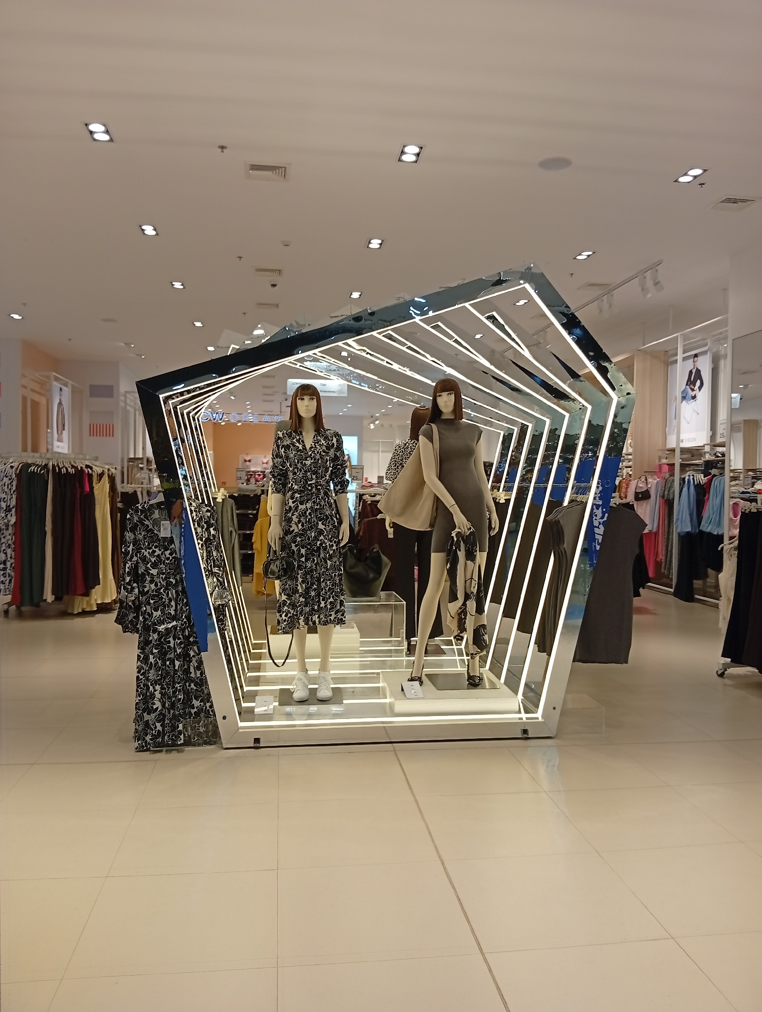 This photograph depicts a retail display, likely inside a department store, set within a well-lit interior space. The focal point is an angular, multi-faceted display structure made with shiny, reflective material and illuminated by linear light strips. Inside this structure, there are two mannequins posed on a raised platform.
The mannequin on the left is wearing a floral print dress and white sneakers, while the one on the right is in a grey dress with black heeled shoes. To the left of the structure, a dress in a similar floral pattern is displayed.
Racks of clothing are visible in the background on both sides of the display, indicating that this is a section within a clothing store. The floor is tiled with large, light-colored squares. The overall impression is that of a clean and organized retail environment. The location is identified as Porlamar, Venezuela.