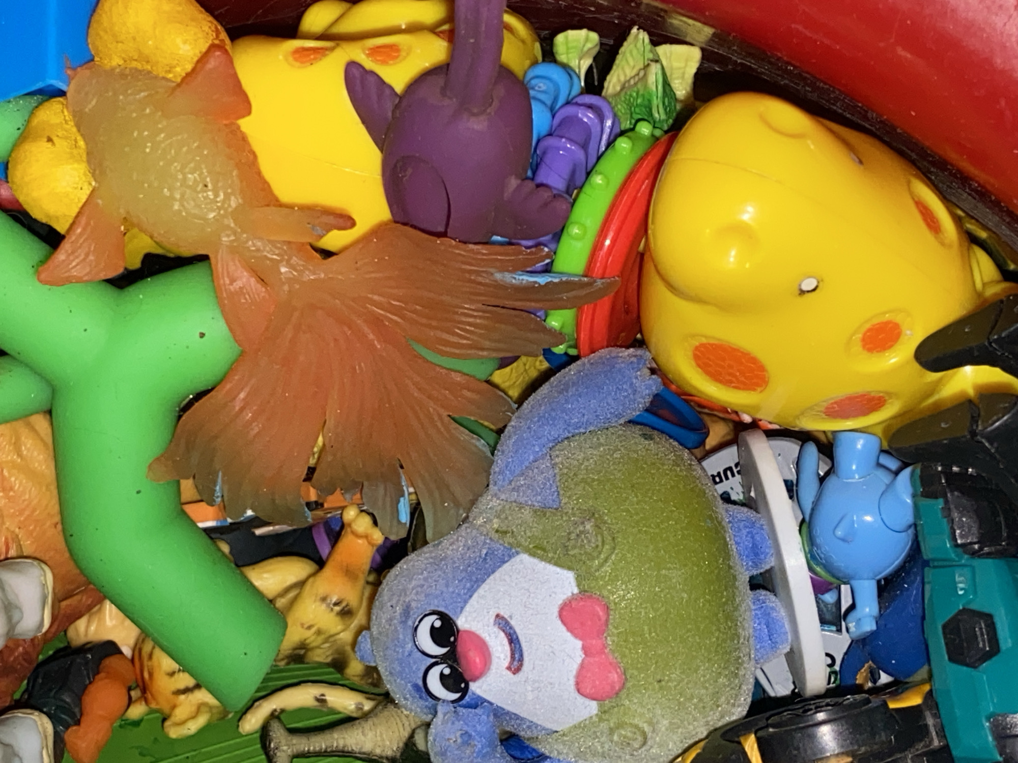 The image depicts a jumble of children's toys of varying shapes, sizes, and colors. Toys include yellow, purple, green, and orange plastic figures. A blue plush toy with a green body is prominently featured in the lower center of the frame. The toys appear to be piled haphazardly in a container. There is no indication that the image was taken in the city of Chaikovsky, Russia.