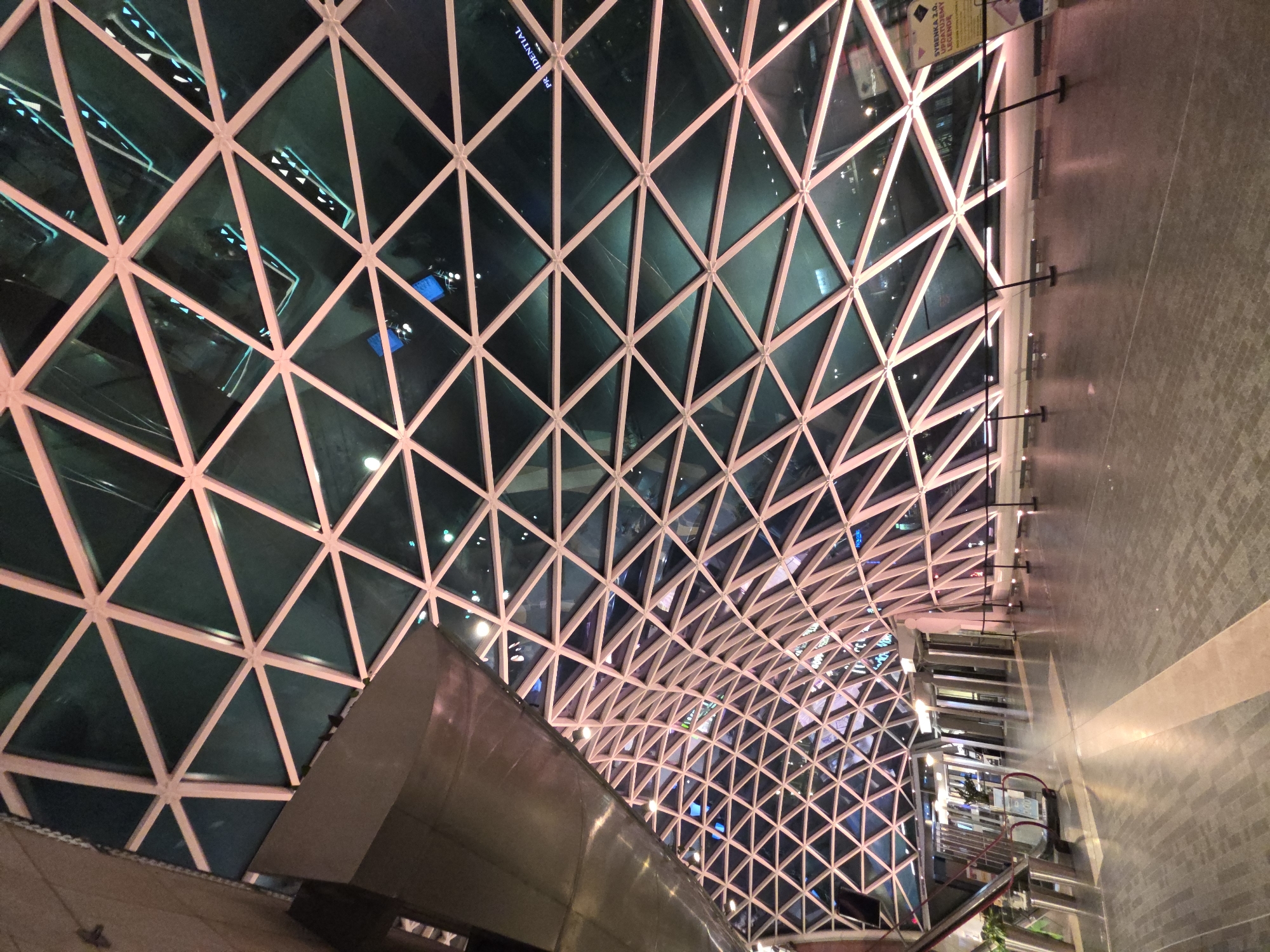 The image depicts the interior of the Złote Tarasy shopping mall in Warsaw, Poland. The most prominent feature is the mall's distinctive glass roof composed of triangular panes supported by a pinkish-beige metal framework. The lighting is dim, and there are reflections of other buildings visible in the glass panes.
In the foreground, there is a segment of a metallic structure, likely part of the mall's architecture. The floor appears to be made of large grey tiles with light reflecting off the surface. Further into the scene, an escalator is visible with the metallic railings.