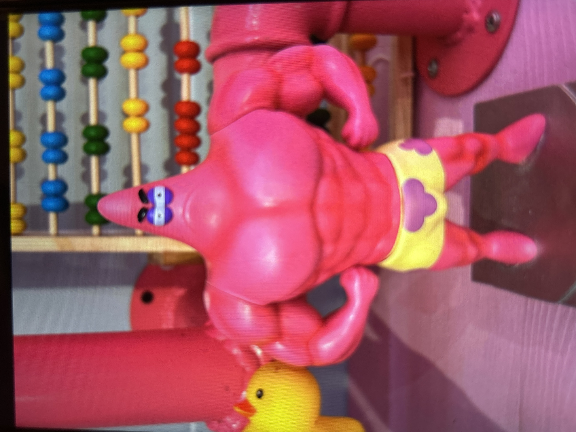 The image features a toy resembling a muscular, anthropomorphic starfish, which appears to be Patrick Star from the Spongebob Squarepants franchise. The toy is predominantly pink and exhibits exaggerated musculature, including prominent biceps and pectorals. It is wearing yellow swim trunks adorned with purple flower-like patterns. The toy is positioned in a vertical orientation, with its head at the bottom and feet pointing upwards.
Behind the toy, a colorful abacus-like structure with rows of beads in blue, green, yellow, and red is visible. The background also includes elements of a pink structure, possibly part of a playground or play area, with visible screws. A small yellow rubber duck toy is partially visible at the bottom of the frame, peeking out from behind the starfish toy.
The lighting suggests an indoor setting, possibly a playroom or a store, with a soft, diffused light. There are no indications of weather or a specific time of day. The overall impression is that of a playful or humorous display of a popular cartoon character depicted in an unusual, hyper-masculine form. No visible text is present in the image.