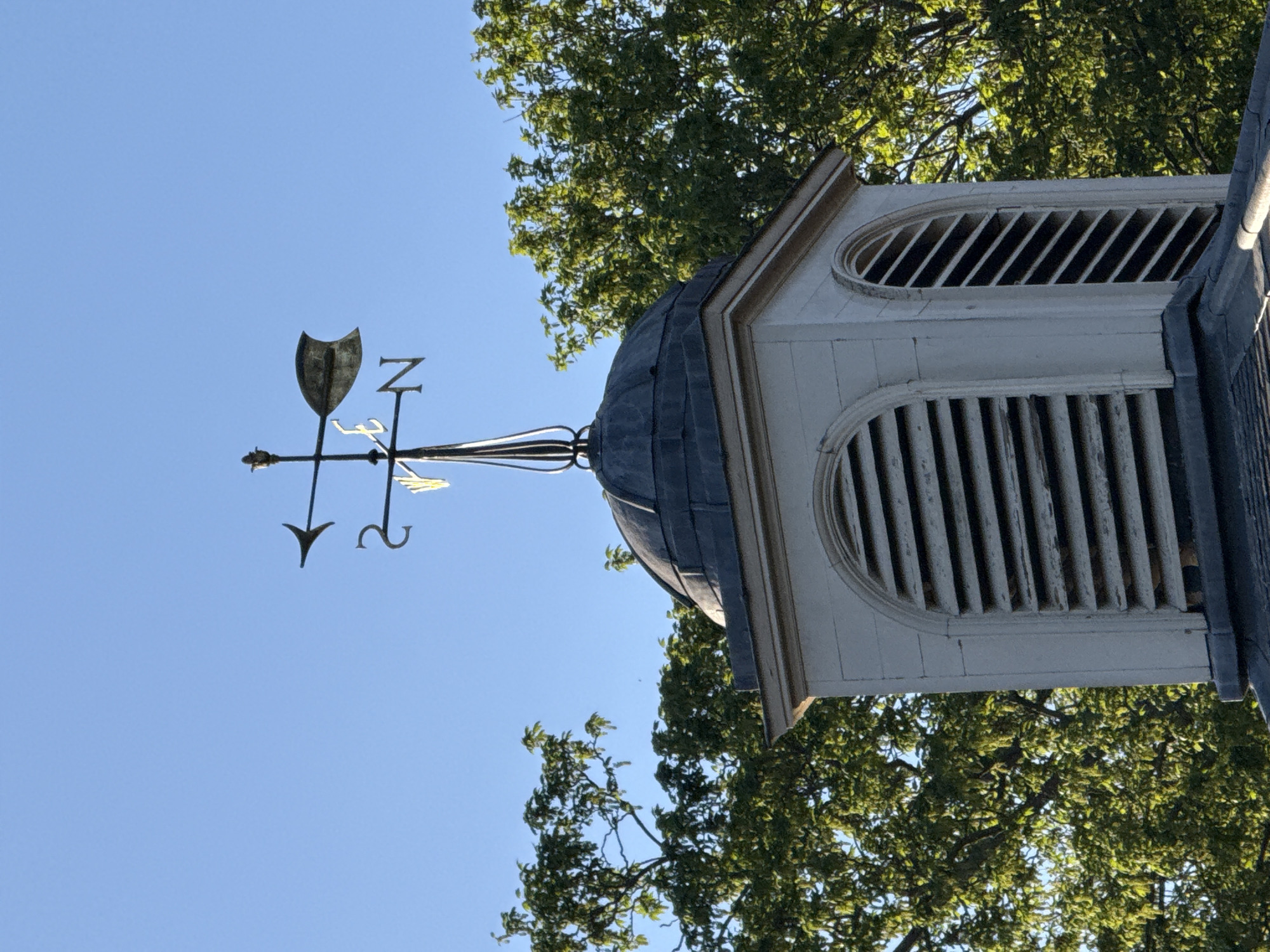 A low-angle view looking up at a small, white-painted cupola with a dark dome and a weather vane on top. The weather vane has an arrow and the letters "N," "E," "S," and "W" indicating the cardinal directions. The cupola has oval-shaped louvered openings on its sides. The scene includes blue sky visible behind the vane and some green trees surrounding the structure. The location is likely the city of London, United Kingdom.