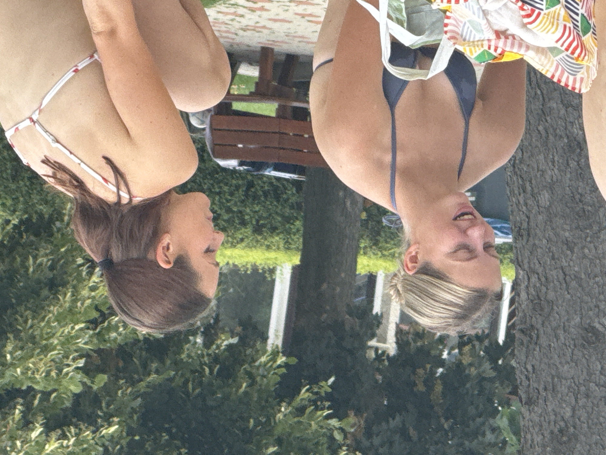 Two young women are sitting outdoors in Hainburg an der Donau, Austria. The woman on the left is facing away from the camera and has her hair pulled back in a bun. She is wearing a dark blue bikini top and appears to be speaking. The woman on the right is facing to the left and has her hair in a ponytail. She is wearing a white bikini top with a red pattern. She is crouching and looking away from the camera. In the background, there is a wooden bench and lush green foliage. The ground is paved with reddish and white tiles. A large tree trunk is visible on the left side of the image.