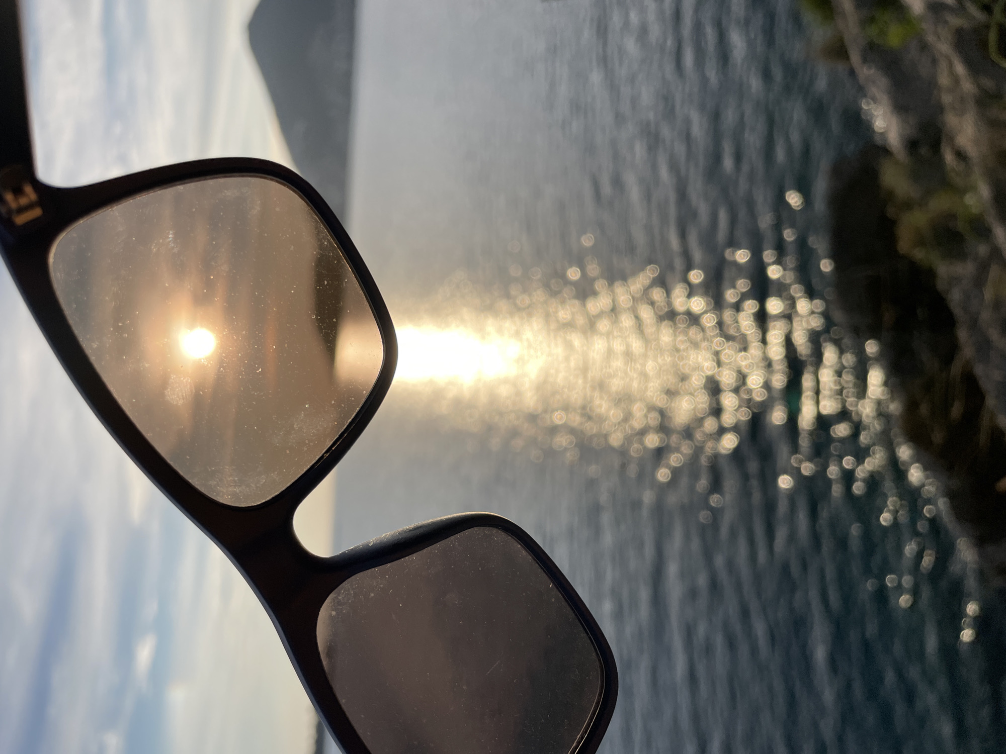 A pair of dark-framed sunglasses is held in the upper central foreground, partially obscuring the background scene. The right lens of the sunglasses exhibits a bright, circular reflection of the sun, which appears orange-yellow, and both lenses show visible dust and smudges. A faint "E" logo is present on the upper right portion of the sunglasses frame.
Beyond the sunglasses, a large body of water, identified as the Adriatic Sea, stretches across the mid-ground, with a path of intense sun reflection creating a shimmering, bokeh effect from the horizon towards the bottom right. The sky above the sea horizon is partly cloudy, displaying a blend of light blue and white tones. A dark, triangular landmass, consistent with a mountain or large hill, is visible on the right side of the frame, silhouetted against the sky. The immediate foreground on the bottom right reveals dark, rugged rock formations with sparse, out-of-focus green vegetation. The scene depicts a coastal environment at either sunrise or sunset, specifically located in Budva, Montenegro, likely from an elevated viewpoint such as a cliff path or lookout point overlooking the sea.
