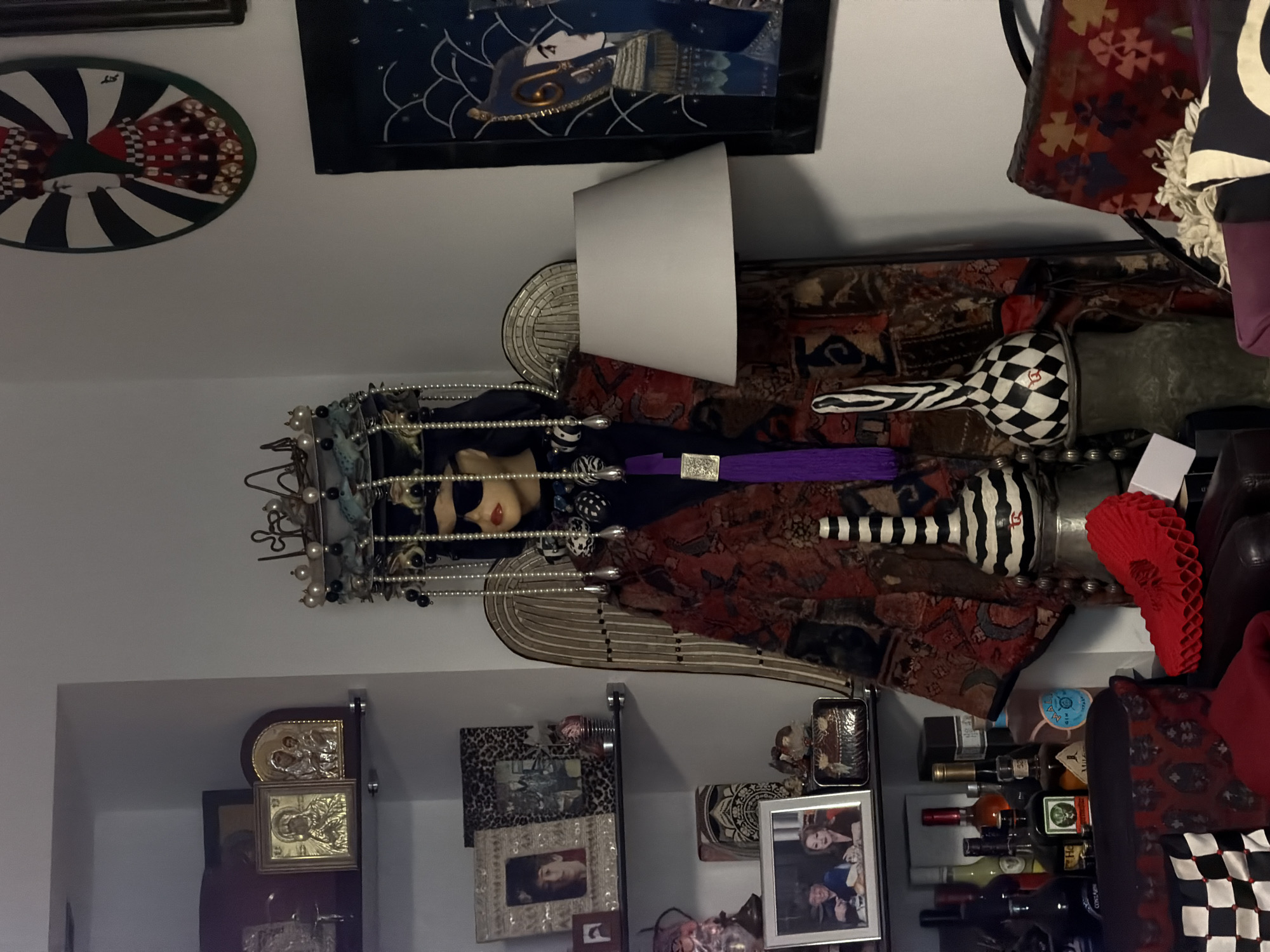 A collection of objects are arranged on shelves. An ornamented figurine is near a lamp with a white shade. Shelves contain decorative boxes, framed pictures, and liquor bottles. Artwork and other decorative items are mounted on the wall. The objects are arranged in a seemingly domestic environment. Location data is not possible to determine from the visual data.