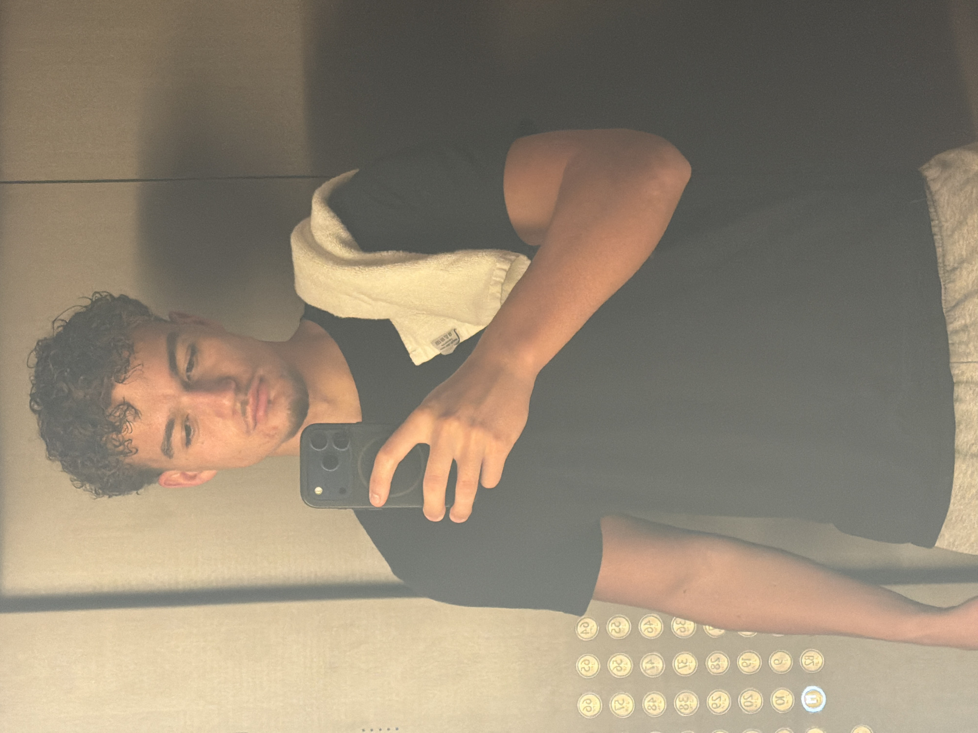 A man with curly hair is taking a selfie in an elevator. He is wearing a black t-shirt and has a white towel draped around his neck. The elevator has a control panel with floor numbers. Based on the reflection and general style, the location is presumed to be within a modern building in Dubai, United Arab Emirates, though definitive proof is absent.