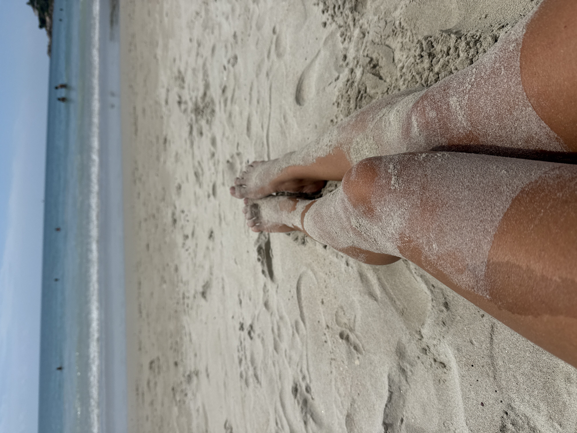 The image captures a close-up view of a person's legs and feet covered in sand, resting on a sandy beach. The legs are tanned and exhibit a tan line on the right thigh. The background shows the calm ocean with gentle waves and a clear blue sky. In the distance, a few people are visible in the water. The scene is likely from a beach in Ko Samui District, Thailand, given the tropical setting. The overall impression is one of relaxation and leisure.