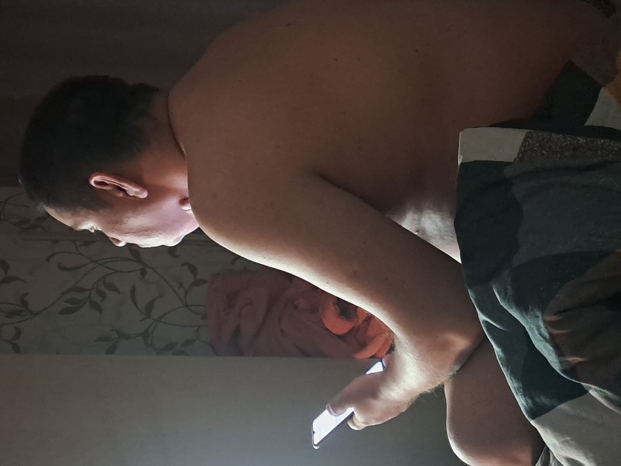The image, rotated 90 degrees clockwise, captures a person intently focused on a smartphone in a dimly lit indoor setting, likely a bedroom. The subject, seen from a high angle, has short, dark hair and fair skin, with their upper body appearing shirtless or minimally clothed.
Their face, neck, and right arm are illuminated by the bright, cool-toned glow emanating from the smartphone screen held in their right hand, indicating active engagement with the device. The light from the phone is the primary light source, strongly suggesting it is nighttime or the room is otherwise dark.
In the background, a wall is visible with a decorative wallpaper featuring a dark, leafy vine pattern. Below the person's arm, a crumpled pile of orange-red fabric, possibly clothing or a blanket, rests against the wall. Dark bedding with light-colored stripes partially covers the person and extends across the right side of the frame. The person appears deeply absorbed in their phone, with no other strong emotions discernible. There is no visible text in the image. The provided location context for this scene is Elektrostal, Russia.