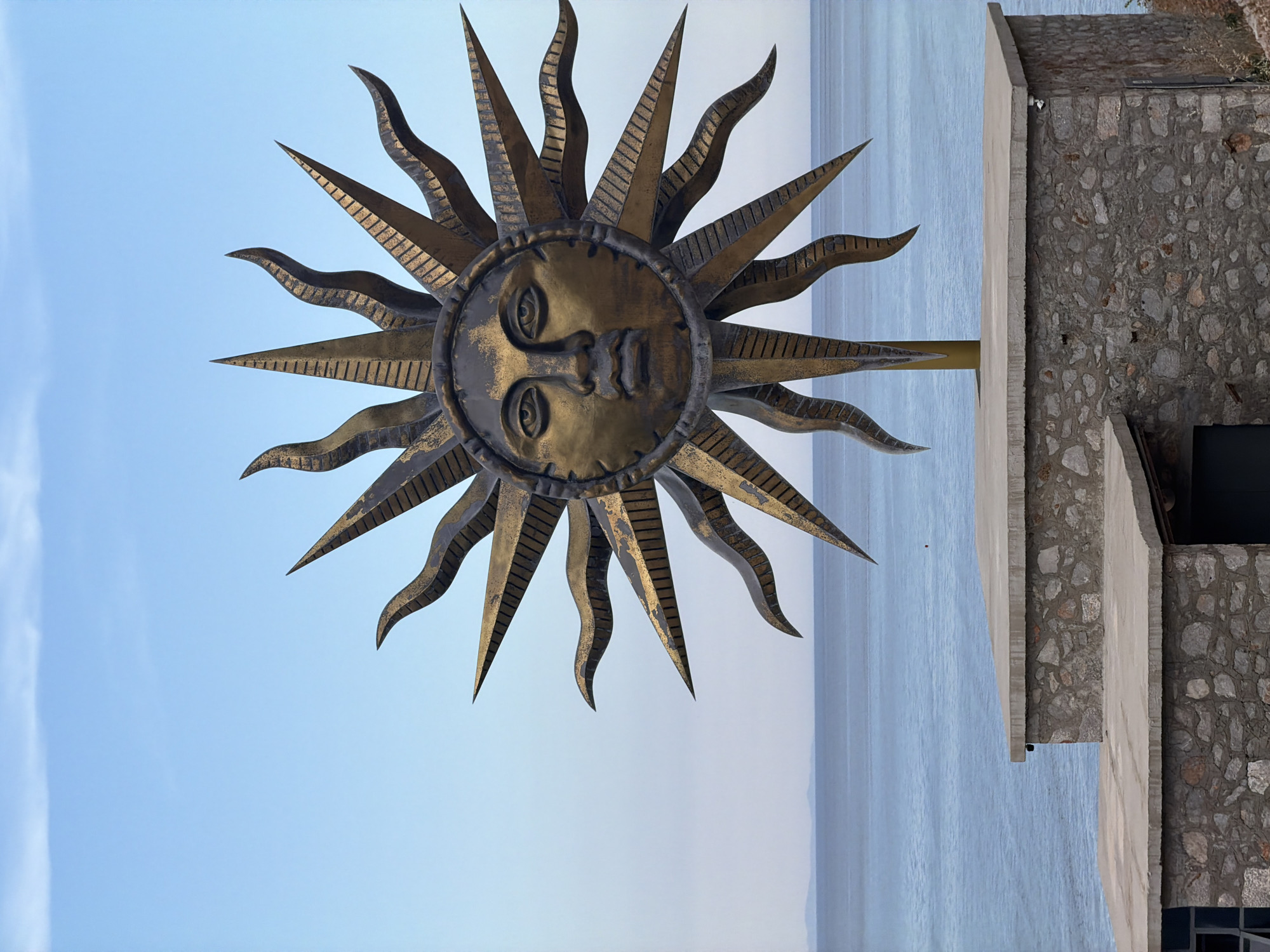 The image depicts a large metal sculpture of a sun with a human face at Hydra Island in Greece. The sculpture is mounted on a metal pole, which is attached to a stone structure. The background consists of a calm sea under a clear, blue sky. The stone structure appears to be a part of a building. The sculpture is the main subject of the image, with its detailed design and intricate metalwork being the focus. The overall environment is scenic.