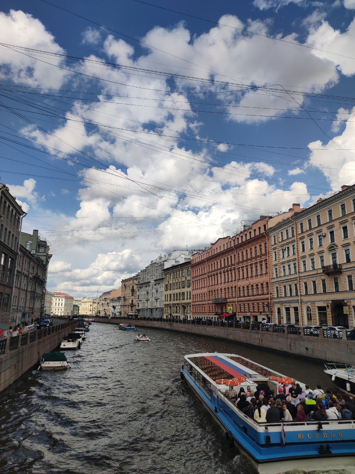 This is a medium-angle, eye-level shot of a canal in Saint Petersburg, Russia, under a cloudy, blue sky. The canal runs horizontally across the image, bordered by stone embankments on either side. A long, blue and white passenger boat, filled with people, is navigating the canal. The boat has a glass enclosure and life preservers are visible along its sides. The buildings lining the canal are tall and multi-story, with a variety of architectural styles and pastel colors, ranging from pale yellow to salmon pink. Several smaller boats are docked or moving along the canal, and cars are visible on the street behind the embankments. Numerous thin wires are strung across the sky, creating a tangled network against the clouds. The overall scene depicts a busy urban waterway with historical architecture and significant human activity.