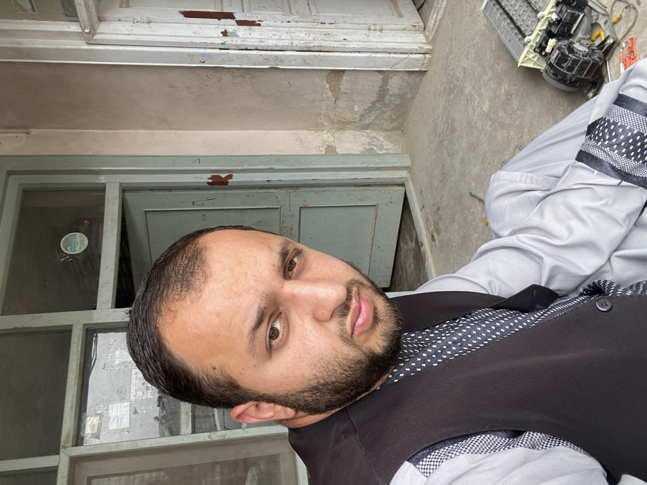 A male individual, appearing to be in his 20s or 30s with dark hair and a full beard, is depicted seated or crouching, facing the camera directly. He is dressed in a light grey long-sleeved garment with patterned cuffs, overlaid by a dark grey waistcoat.
The scene is an outdoor or semi-outdoor area, likely a patio or porch of a weathered building in Kandahar, Afghanistan. Behind the individual, a light blue-green wooden door with multiple glass panels is visible, partially open. A circular sticker is affixed to an upper glass pane. To the right of this door, a pale, possibly pink or beige, wall exhibits significant peeling paint and moisture damage near its base. Further right, a second weathered wooden door frame, originally light-colored with remnants of reddish-brown paint, is present. The ground is a grey concrete surface.
To the lower right of the frame, on the concrete, lie various disassembled electronic components, including multiple rectangular battery modules and a black electric motor or fan unit, alongside what appear to be hand tools such as a screwdriver and assorted wires, suggesting repair or technical work.
