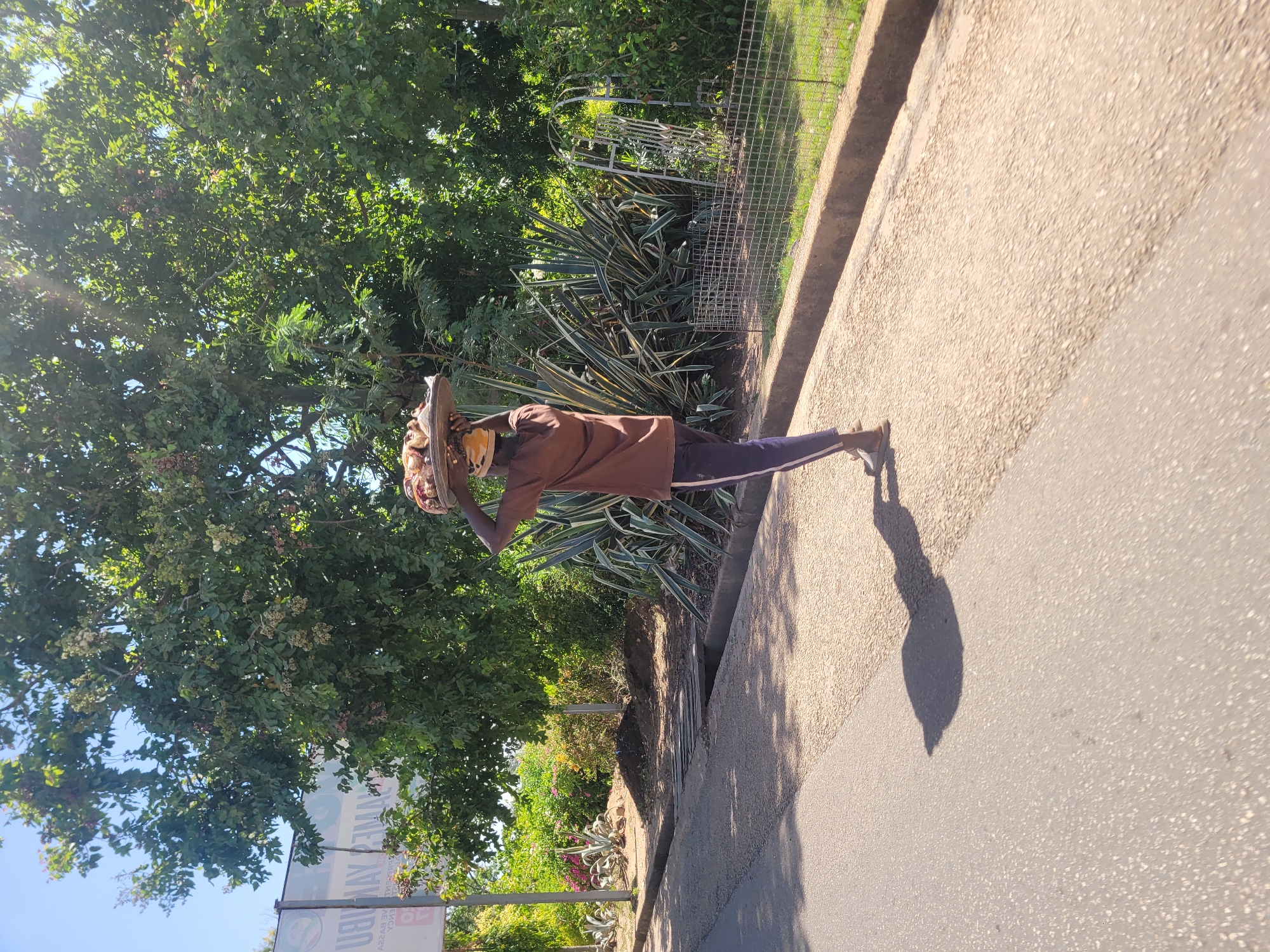 The image shows a person walking along a street in Jos, Nigeria. The person is wearing a brown shirt and dark pants with a white stripe down the side. They are carrying a basket on their head. The street is paved and has a sidewalk. There are trees and bushes along the side of the road. A sign is visible in the bottom left corner of the frame. The sun is shining brightly, casting a shadow of the person on the road.