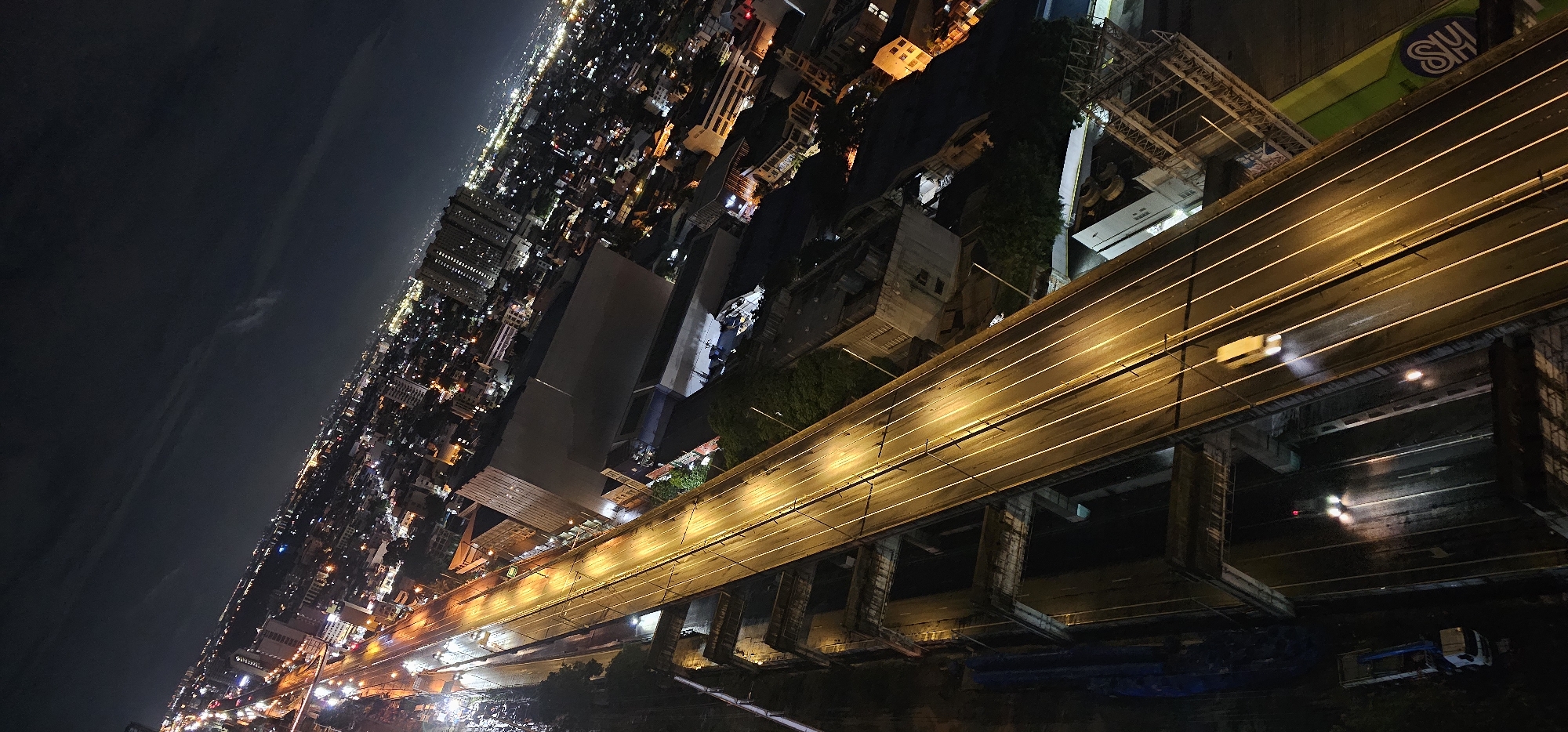 An aerial, high-angle night shot depicts a brightly lit elevated highway cutting through the dense urban landscape of Makati, Philippines. The highway, illuminated by continuous rows of warm yellow lights, is the central focus, with a lone white vehicle traversing its lanes, its headlights creating streaks of light. The highway structure itself is supported by massive concrete pillars, some appearing partially constructed or aged.
Beyond the highway, the city sprawls out in a vast panorama of lights. Countless buildings of varying heights and architectural styles are visible, their windows twinkling with a multitude of colors, predominantly orange, yellow, and white. The distant skyline fades into a hazy glow under the dark night sky, suggesting a sprawling metropolis.
Below the elevated highway, glimpses of the city at ground level are visible. These include what appear to be lower-rise buildings, some with illuminated windows, and patches of trees. In the foreground, the edges of other structures and infrastructure can be seen, including what looks like a truck with a crane attachment on the left and a large green billboard with a blue logo (possibly "SM") on the right. The overall impression is one of a vibrant, bustling city at night, with the elevated highway serving as a prominent artery of movement and light.
