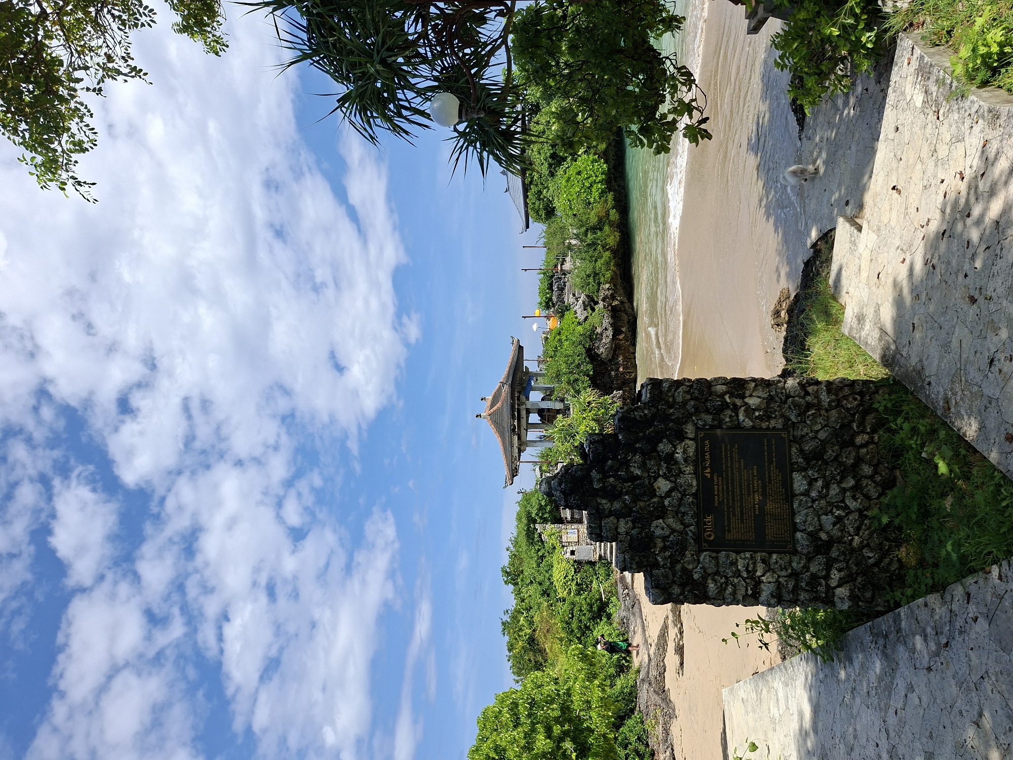 The image captures a view of Nusa Dua Beach in Kuta Selatan, Indonesia. The scene features a sandy beach along a coastline with a structure reminiscent of a traditional Balinese pavilion or temple perched on a cliff overlooking the ocean. Lush tropical vegetation surrounds the building and the coastal area. A small, white dog stands on the shore near some stone steps. In the foreground, a stone structure covered with a plaque is visible. The sky is bright blue with scattered white clouds.