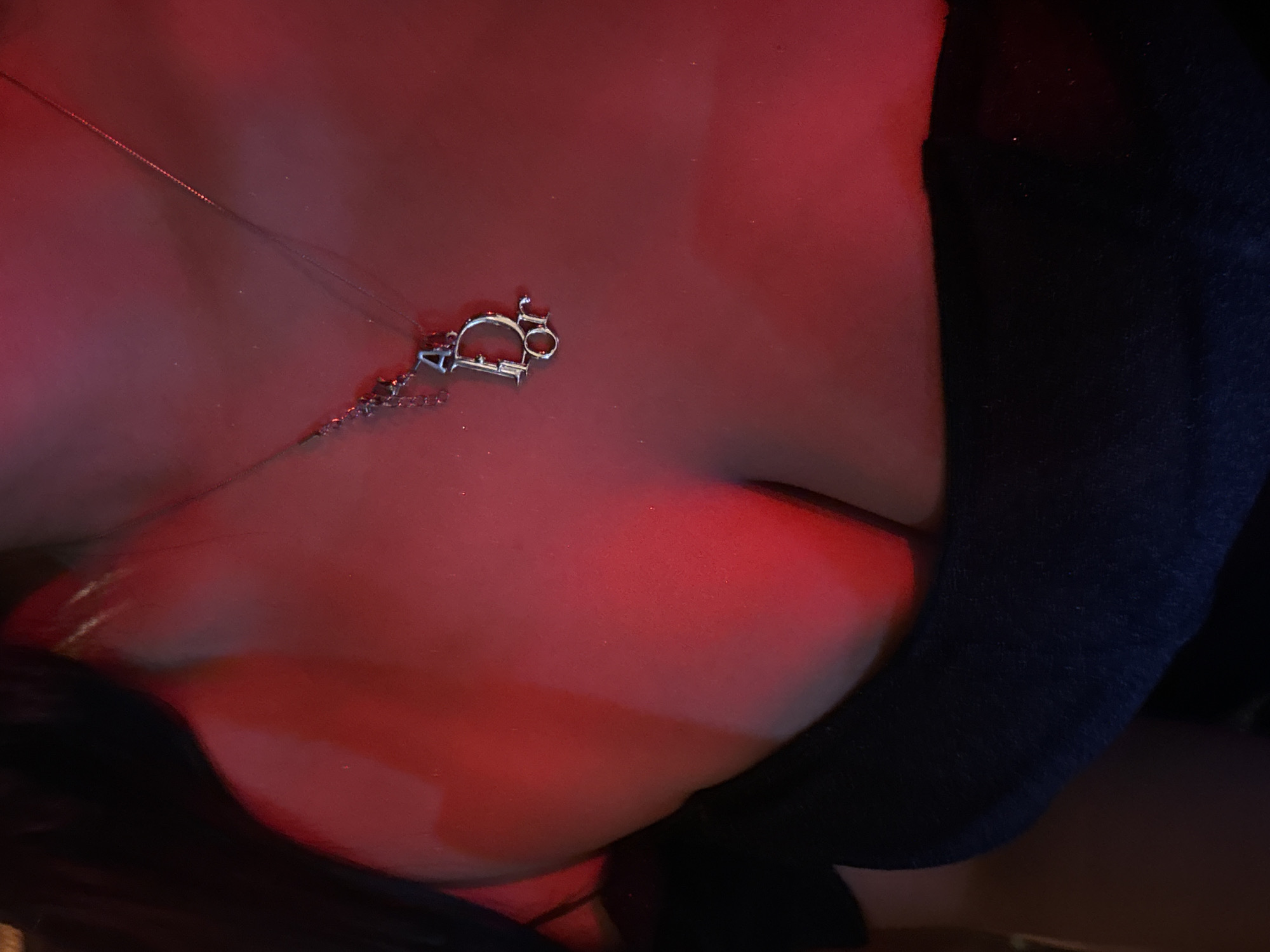 The image shows a close-up of a person's chest wearing a black off-the-shoulder top and a silver Dior necklace. The lighting is predominantly red, creating a dramatic effect. The background is dark and out of focus, with hints of dark hair visible on the left side. The image is taken from a chest-up perspective, focusing on the necklace and the décolletage. No specific location in North Kuta, Indonesia, can be identified from the image itself.