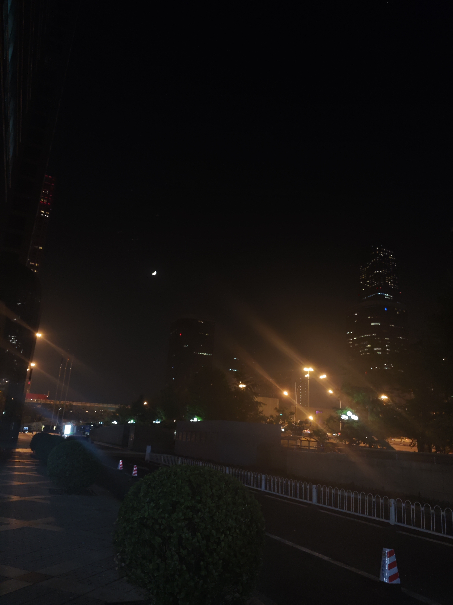 This nighttime image captures a quiet urban street scene, likely in Chaoyang District, China. The dark sky features a distinct crescent moon, positioned slightly left of center. The background is dominated by modern city architecture, including several tall skyscrapers. The most prominent building on the far right is heavily lit with numerous windows, while another, slightly shorter building with scattered lights, stands further left.
In the foreground, a paved path or sidewalk is visible, lined with dark, rounded bushes. To the right of these bushes, a road is discernible, separated by a low white metal fence. Two red and white striped traffic cones are placed on the ground, one in the bottom right corner and another further back. Bright streetlights cast warm yellow and white glows, creating prominent lens flares and illuminating parts of the surrounding environment, including distant trees. A multi-lane bridge or overpass, also lit, spans the mid-ground. The scene is devoid of people, conveying a sense of urban tranquility. No legible text is visible.