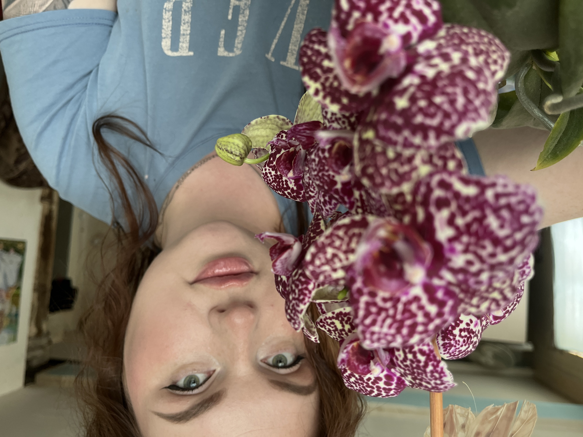 A woman is holding a spotted purple and white orchid close to her face. She has fair skin, light green eyes with dark eyeliner, and light pink lips. Her auburn hair is pulled back. She is wearing a light blue t-shirt with white lettering. The background appears to be an interior space. The location is Павловка, Russia.