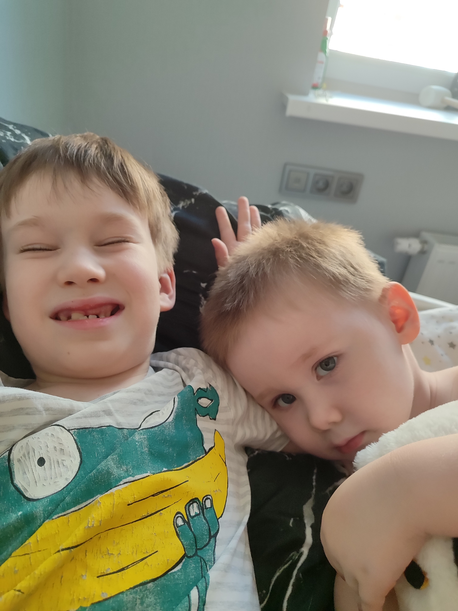 Two young children are lying down indoors. The older child on the left is smiling with his eyes closed, and he is missing some front teeth. He is wearing a gray and white striped shirt with a faded green and yellow cartoon graphic. The younger child on the right is looking at the camera with a thoughtful expression and holding a stuffed animal. His hand is raised with two fingers extended. They appear to be lying on a bed with black and white patterned sheets. Behind them is a window with a white frame. The scene is brightly lit. It is not possible to ascertain that this image was taken in Izhevsk, Russia.