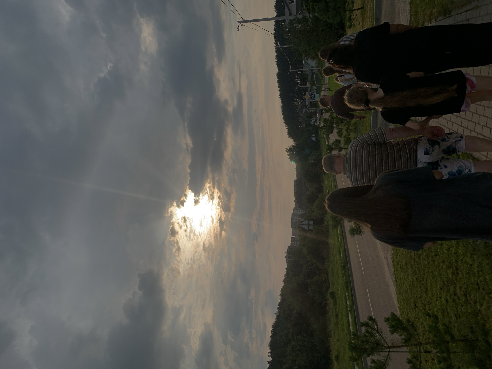 A group of people walk on a paved path next to a road. The sky is overcast with the sun breaking through the clouds, creating visible sunbeams. In the distance, there are buildings and trees. The people are walking away from the camera. One person in the foreground is wearing a dark shirt and has long brown hair. Behind them, a person in a striped shirt and patterned shorts walks with others. A young girl with long blonde hair in a ponytail walks near the center, holding hands with an adult. Further back, more people are visible, including a person in a black shirt and dark pants. The environment appears to be a suburban or semi-rural area.