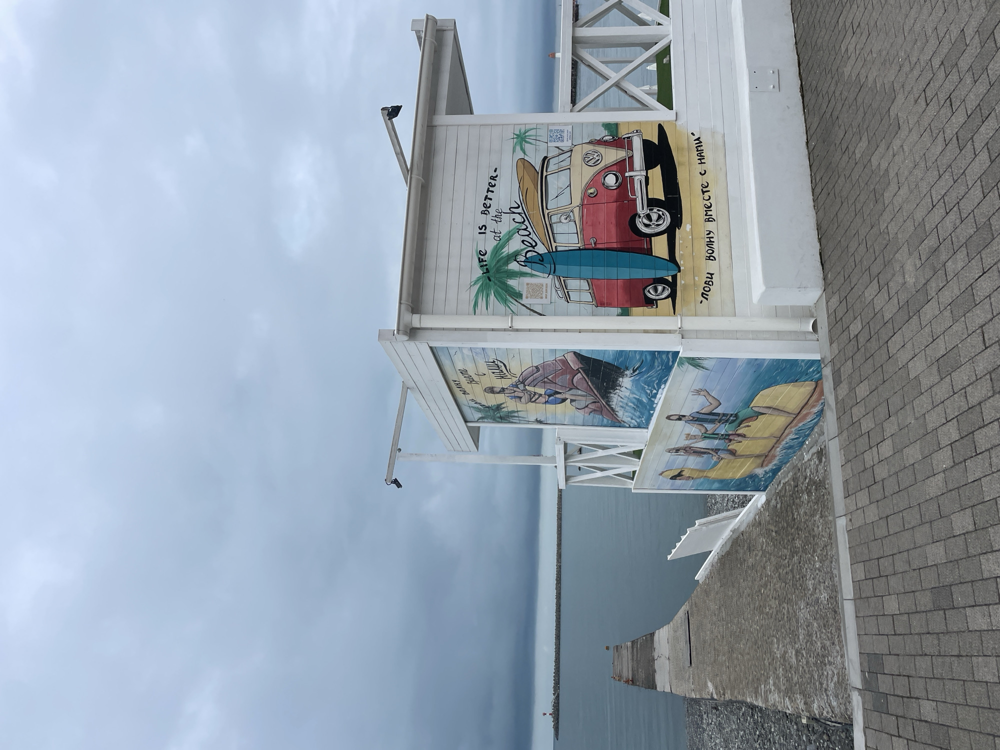The image shows a beachside structure with murals, situated at a location that appears to be in Sochi, Russia, based on the Cyrillic script on the murals. The structure is white, with multiple levels of murals depicting beach scenes. The top mural features a red and beige VW van with surfboards. The phrase "Life is better at the beach" is written in English, along with "Лови волну вместе с нами," a Russian phrase meaning "Catch the wave with us." The lower murals show people engaged in water activities, such as surfing or boating. The structure is positioned along a paved walkway next to a concrete seawall or pier. Beyond the pier, there is a view of the sea and a cloudy sky. The scene has a beachside or coastal atmosphere.