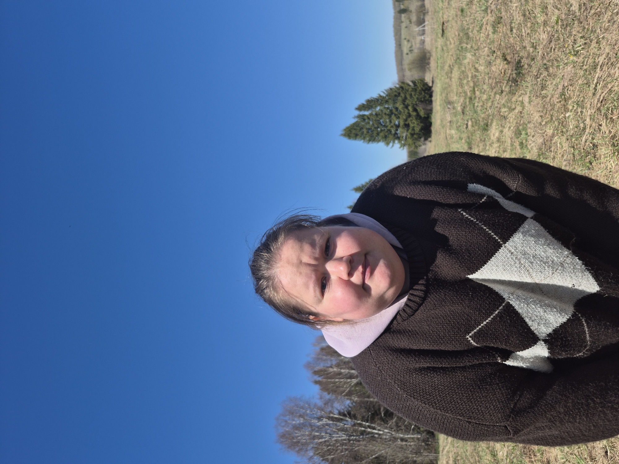 A person is in a field, possibly in Дойная, Russia. The person is wearing a brown sweater with a white argyle pattern. The sky is blue and clear. The ground is covered in dry grass and some green grass. Some bare trees are in the background.