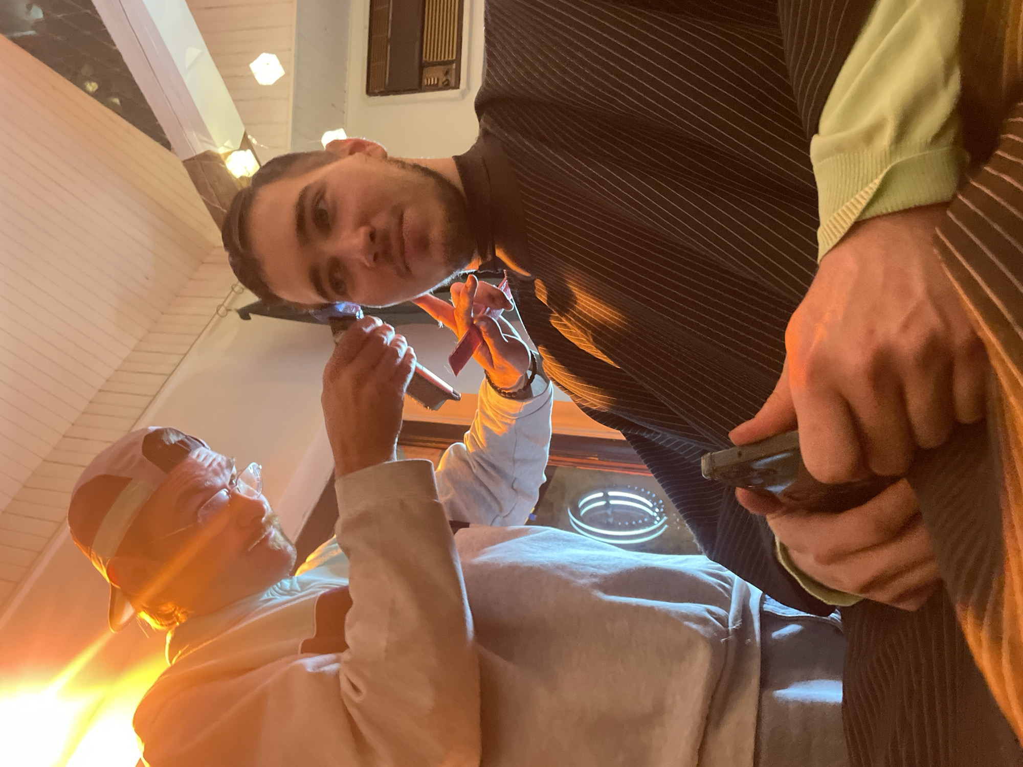 Two men are in an indoor location. One man wears a baseball cap, glasses, and a light-colored hoodie. He holds a small device. The other man wears a dark pinstripe suit, light-green shirt, and holds a smartphone. A partially visible illuminated decorative object and ceiling lights are present. The location is Belgrade, Serbia.