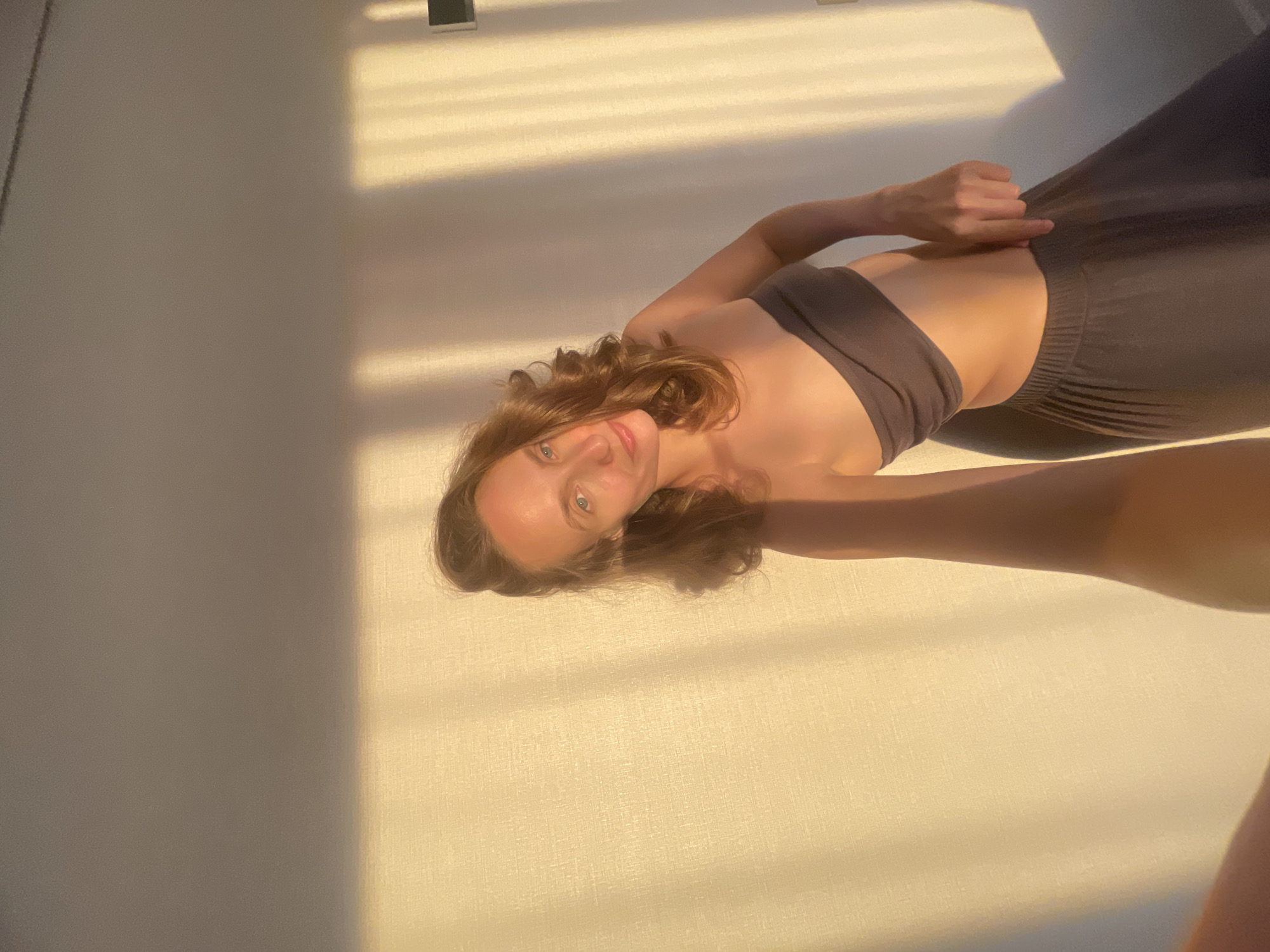 A woman is captured in a selfie taken indoors. She is wearing a dark strapless top and a dark, flowing skirt. The room appears to be lit by natural light, casting elongated shadows on the wall behind her, suggesting it might be late afternoon or early morning. The wall has a textured surface, possibly wallpaper or a stucco finish. The overall mood is relaxed and casual. The image does not provide any specific details that would allow for the identification of the location within the city of Подшивалово, Russia.