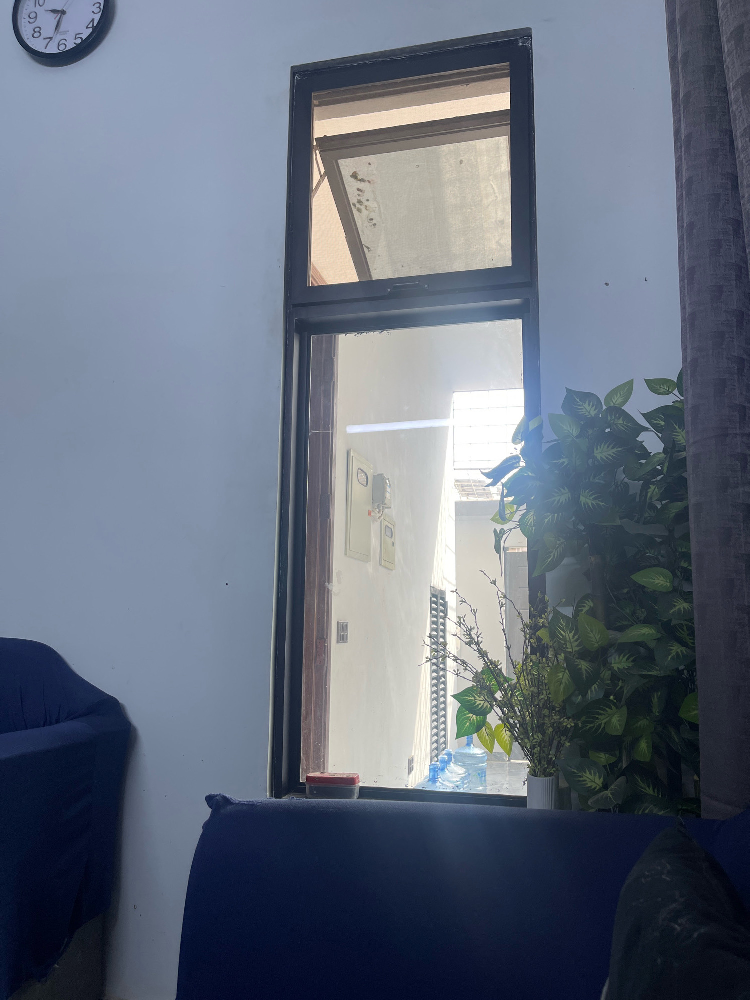 The image is an interior shot of a room, looking out a tall, narrow window. The wall to the left is a pale blue. A black and white analog clock with Roman numerals is visible on the upper left portion of the wall. To the right of the clock, and filling most of the frame, is the window. Through the window, an outdoor scene is visible, characterized by bright sunlight and architectural elements including what appears to be a building with white walls and some metal grating. Several large blue water bottles are positioned on a ledge outside the window. A large, leafy green artificial plant is situated to the right of the window, partially obscuring the view. A dark grey curtain hangs to the right of the window. In the foreground, parts of blue upholstered furniture are visible, suggesting this is a living space. There are no people or animals in the frame, and no explicit action is taking place. The scene suggests a domestic environment in Karachi Division, Pakistan, as inferred by the presence of large water bottles, a common sight in areas with inconsistent municipal water supply.