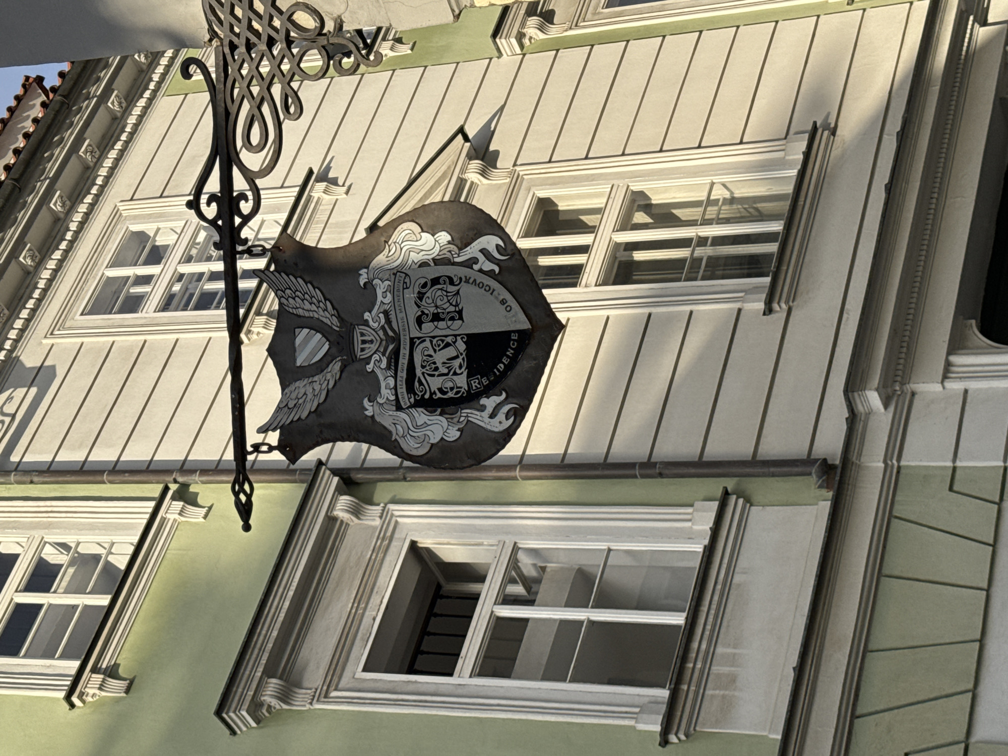 An outdoor, eye-level medium shot shows a building facade in Prague, Czechia, on a sunny day. The building is made of light beige and green painted stucco, with white trim around the windows and architectural details. A wrought-iron bracket hangs from the building, supporting a dark shield with an emblem and text. The shield is decorated with wings and a coat of arms divided into four sections, with ornate scrollwork and Latin text. Two windows are visible on the light beige section of the building, each with a white frame and multiple panes. The lower part of the building is painted a light green, also with white trim. The sun casts shadows across the facade, highlighting the textured surfaces and architectural features.