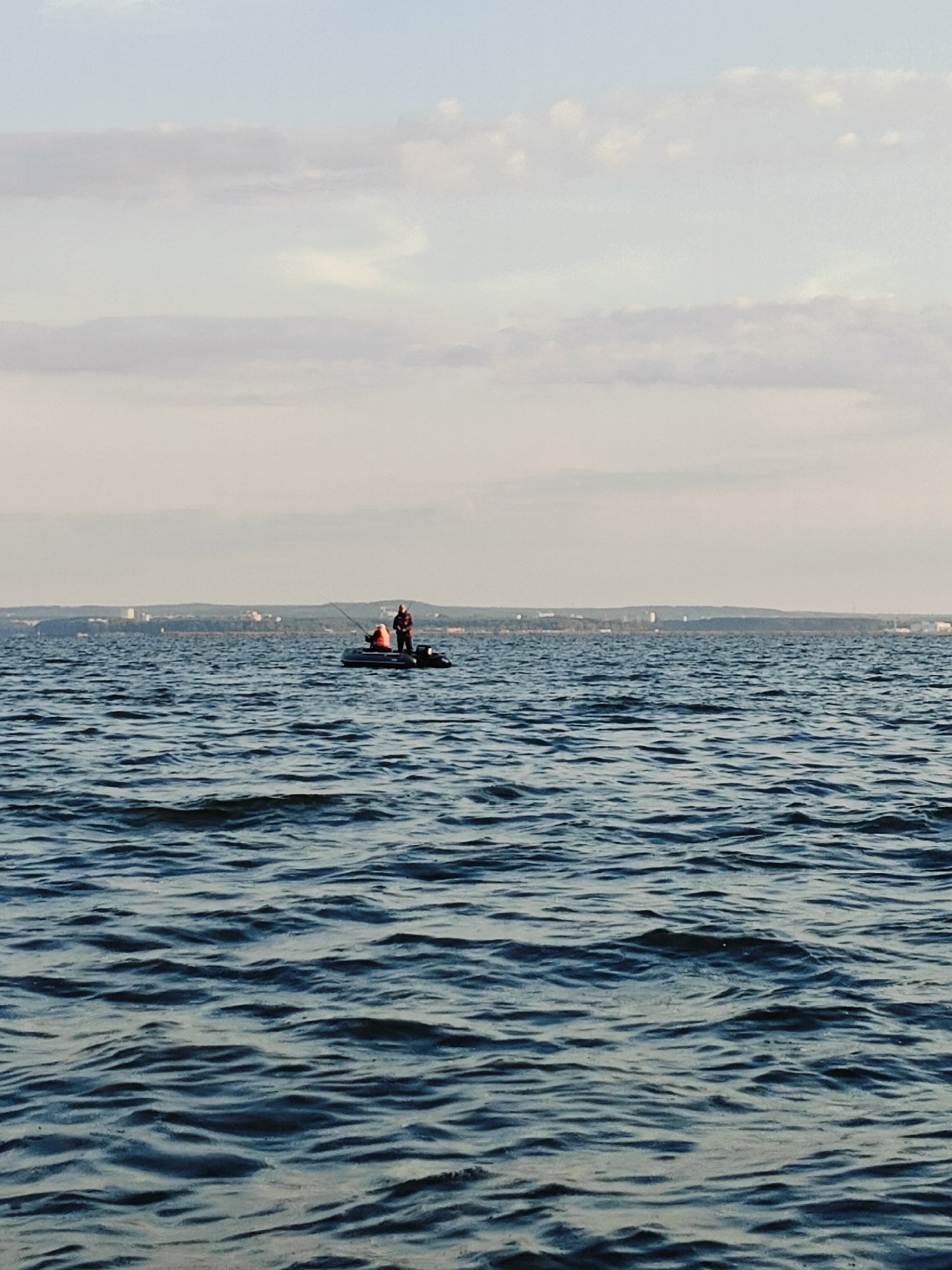 The image depicts a tranquil scene on the Kama River near Chaykovsky, Russia. In the foreground, gentle blue waves ripple across the water's surface. In the mid-ground, a small inflatable boat carries two individuals, one of whom appears to be fishing with a rod. The boat is positioned on the calm river, with a distant, hazy shoreline in the background. The shoreline features a band of trees and structures, suggesting the presence of a town or city. The sky above is a pale blue, adorned with streaks of white clouds. The overall impression is one of peaceful outdoor activity and natural beauty.