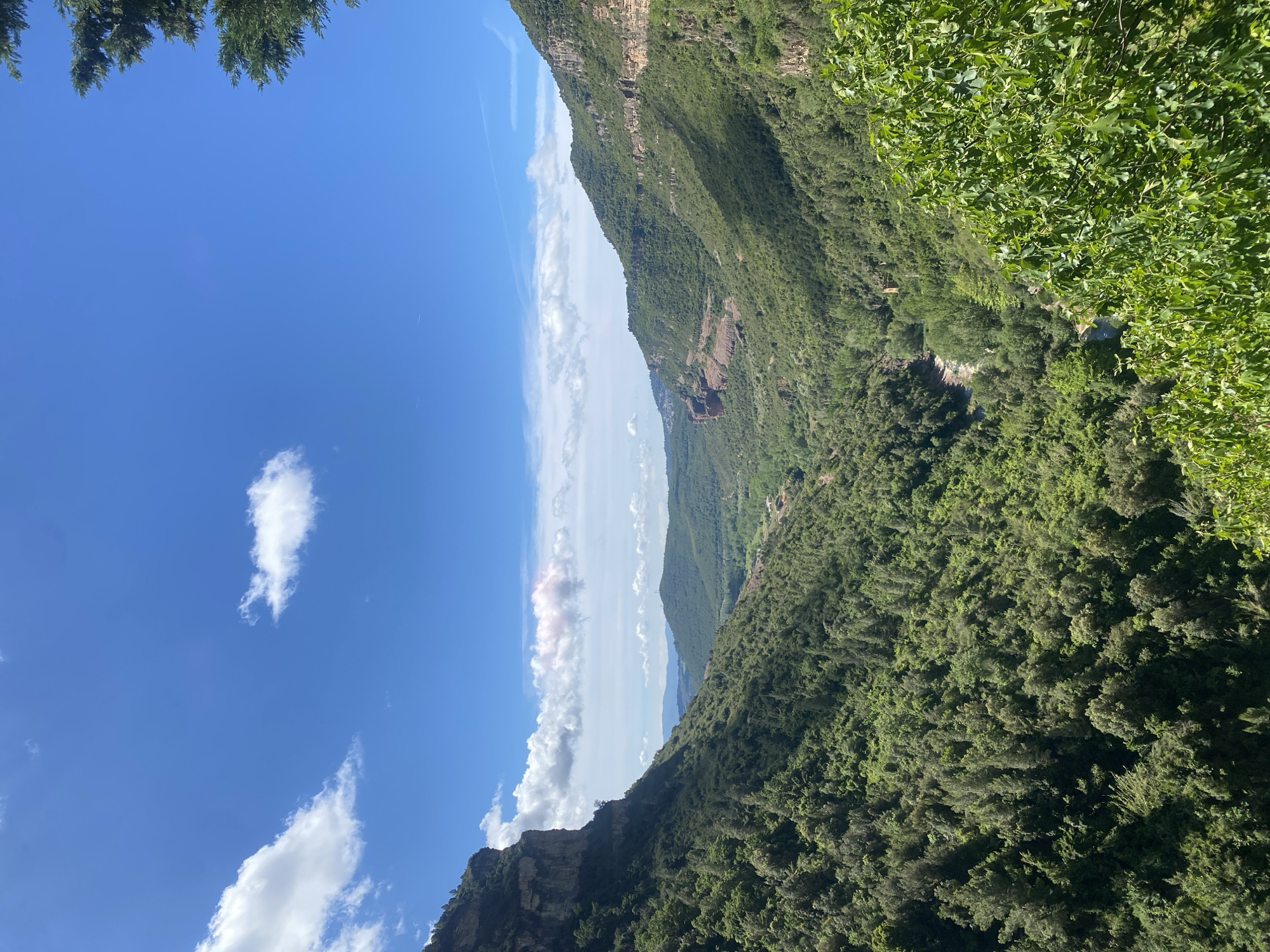 This is an exterior shot taken at an upward angle in what appears to be a mountainous region. The scene features a deep canyon or valley with steep, densely forested slopes. The foliage is lush and green, suggesting a moist or temperate climate. The sky is predominantly blue, with scattered white clouds. A significant geological feature is a prominent rock formation with visible layers, suggesting sedimentary rock. The image location is Sant Quirze Safaja, Spain.
