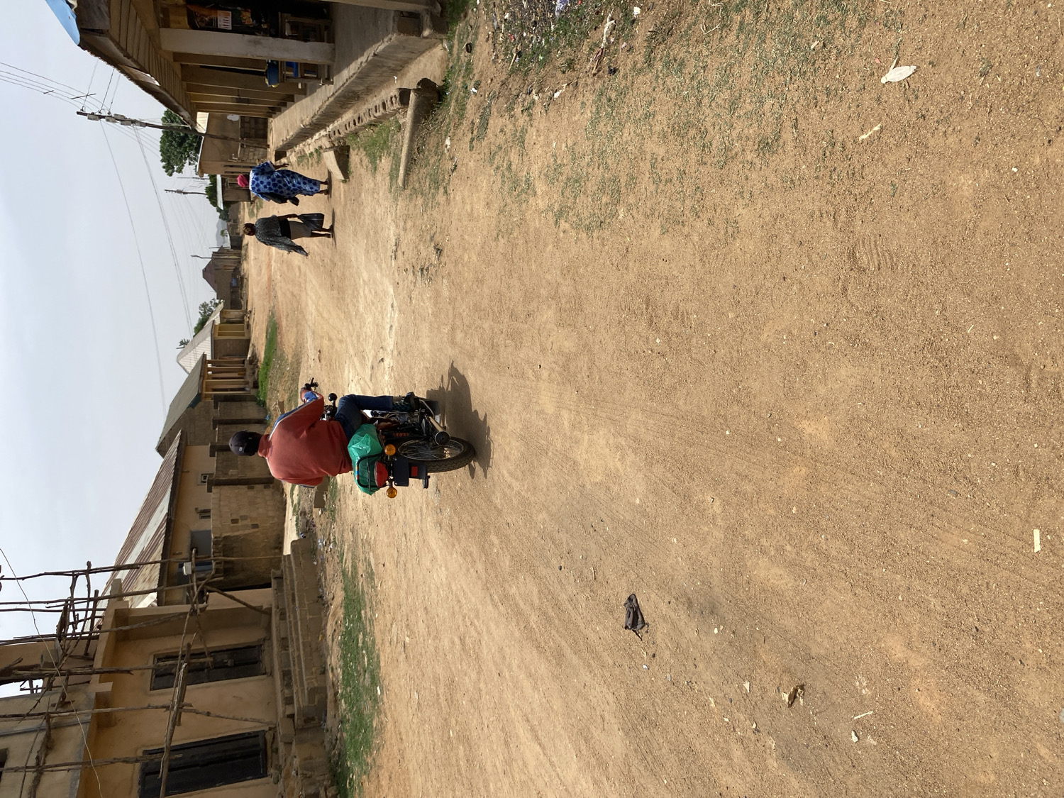 The image captures a street scene in Barakin Akawo, Nigeria, seen from a slightly elevated perspective. The main subject is a man riding a motorcycle away from the viewer, positioned in the lower-left to central part of the frame. He is wearing a red long-sleeved shirt, blue jeans, and a dark helmet, with a green bag strapped to the back of his motorcycle.
Further up the dirt road, three individuals are walking. Two women are more clearly visible: one in a vibrant blue and white patterned outfit with a headscarf, and another in a darker, possibly grey or black, patterned dress. A third person is walking behind them, partially obscured.
The setting is characterized by a wide, unpaved dirt road that stretches into the distance, showing visible tire tracks. On both sides of the road, various single-story buildings line the street. Some buildings appear to be residential or commercial structures, with corrugated metal roofs. On the left side, closer to the foreground, there's a building under construction, with wooden scaffolding. Utility poles with overhead wires run alongside the road. The ground along the edges of the road shows patches of sparse green grass and some debris.
The sky appears clear and bright, indicating daytime. The strong sunlight casts shadows, suggesting a mid-morning or afternoon time. No discernible text is visible on any of the buildings or signs. The overall impression is one of a busy, semi-urban or rural environment during daily activity.
