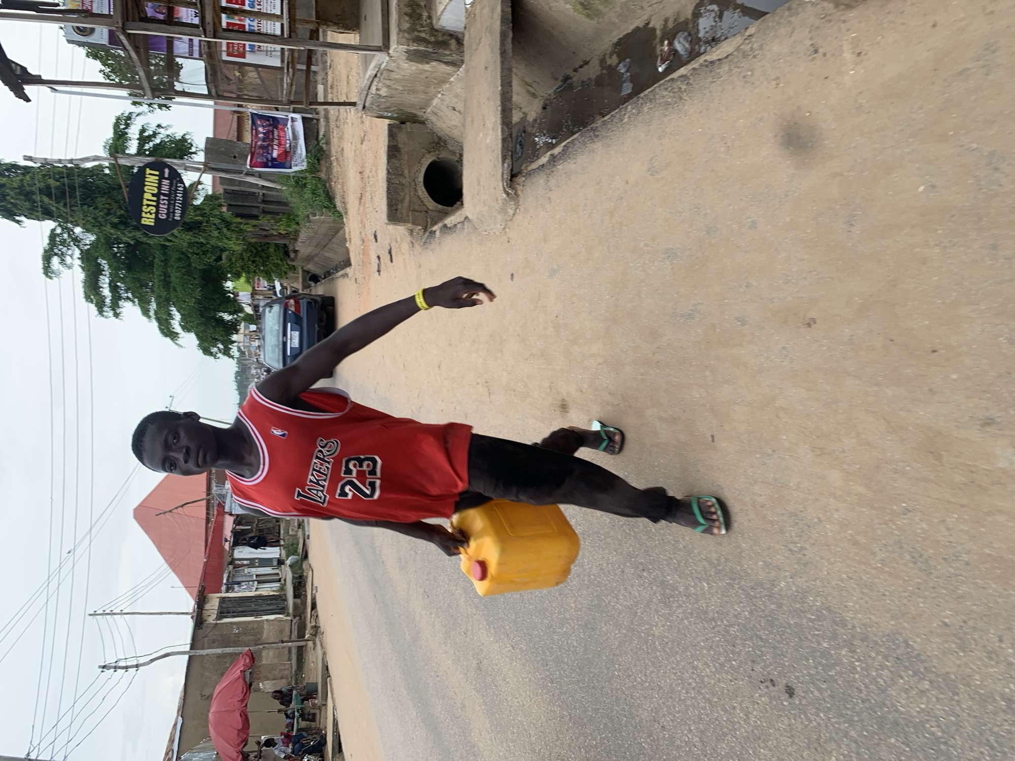 A young Black male is walking on a paved road in Abuja, Nigeria, carrying a bright yellow plastic jerrycan in his left hand. He is wearing a red basketball jersey with "LAKERS" and the number "23" in white and black lettering, black pants, and green flip-flops. He has a yellow wristband on his right wrist. The road appears to be a commercial street with various stalls and buildings lining one side, including a red umbrella providing shade for some people sitting at a table. In the background, there is a tall green tree and a sign that reads "RESTPOINT GUEST INN" with a phone number. A dark-colored car is visible on the road. The ground is a mix of paved surfaces and dirt, with a concrete drainage channel running along the side of the road. The sky is overcast.